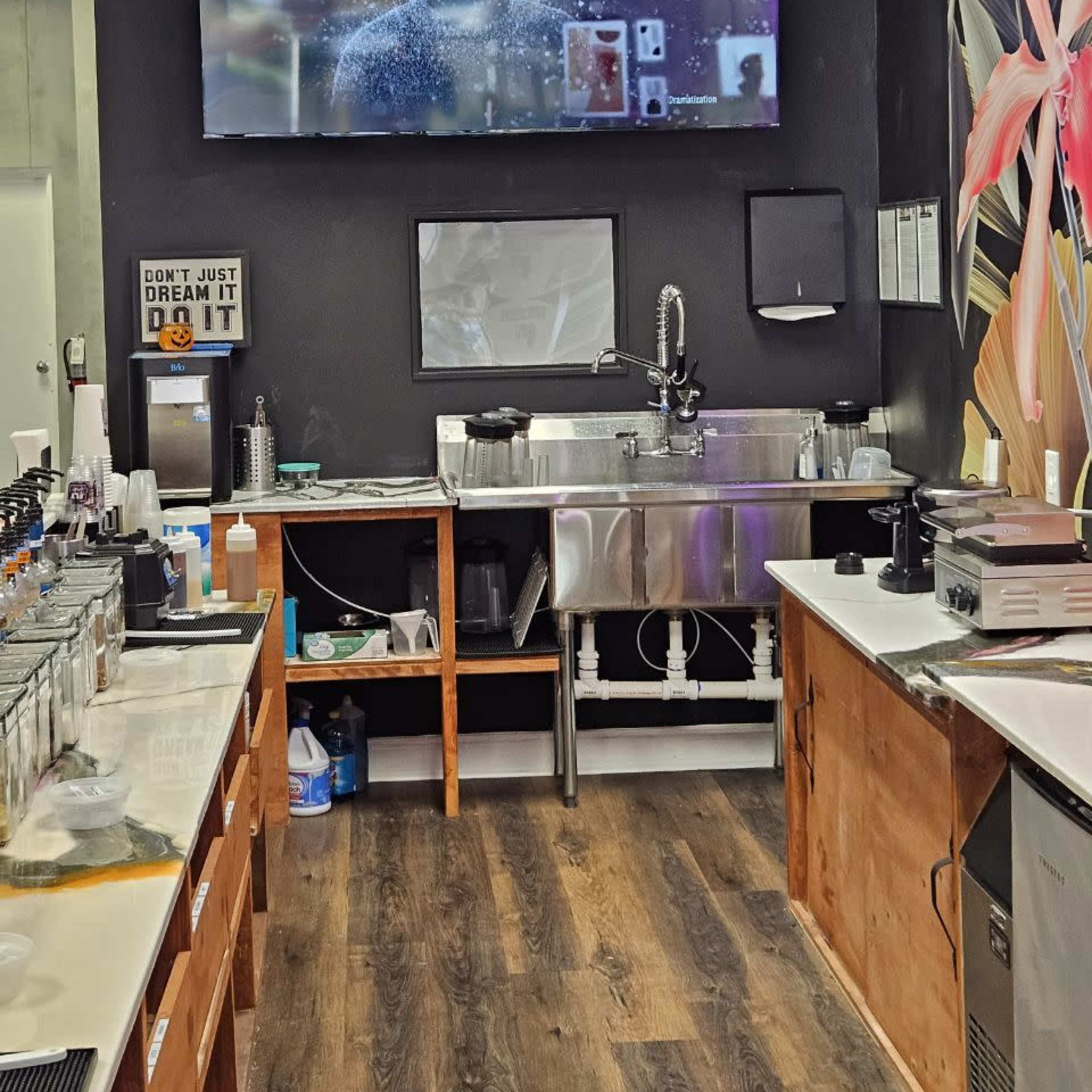 A café interior featuring a large television on the wall displaying a digital graphic, along with a sink and work counter with various glassware and equipment.