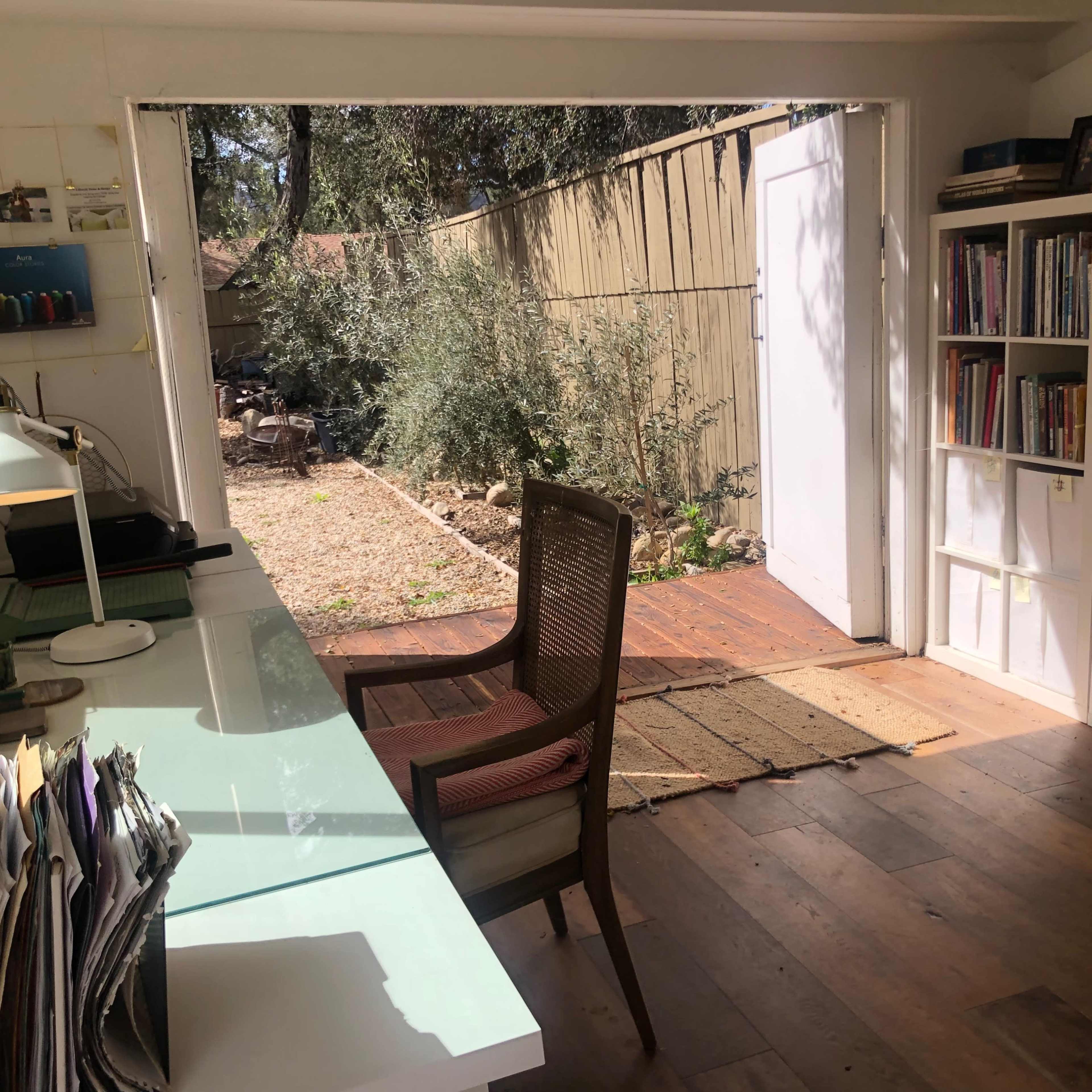 A room with a desk and chair facing an open door that leads to a garden area, with bookshelves filled with books along one wall.