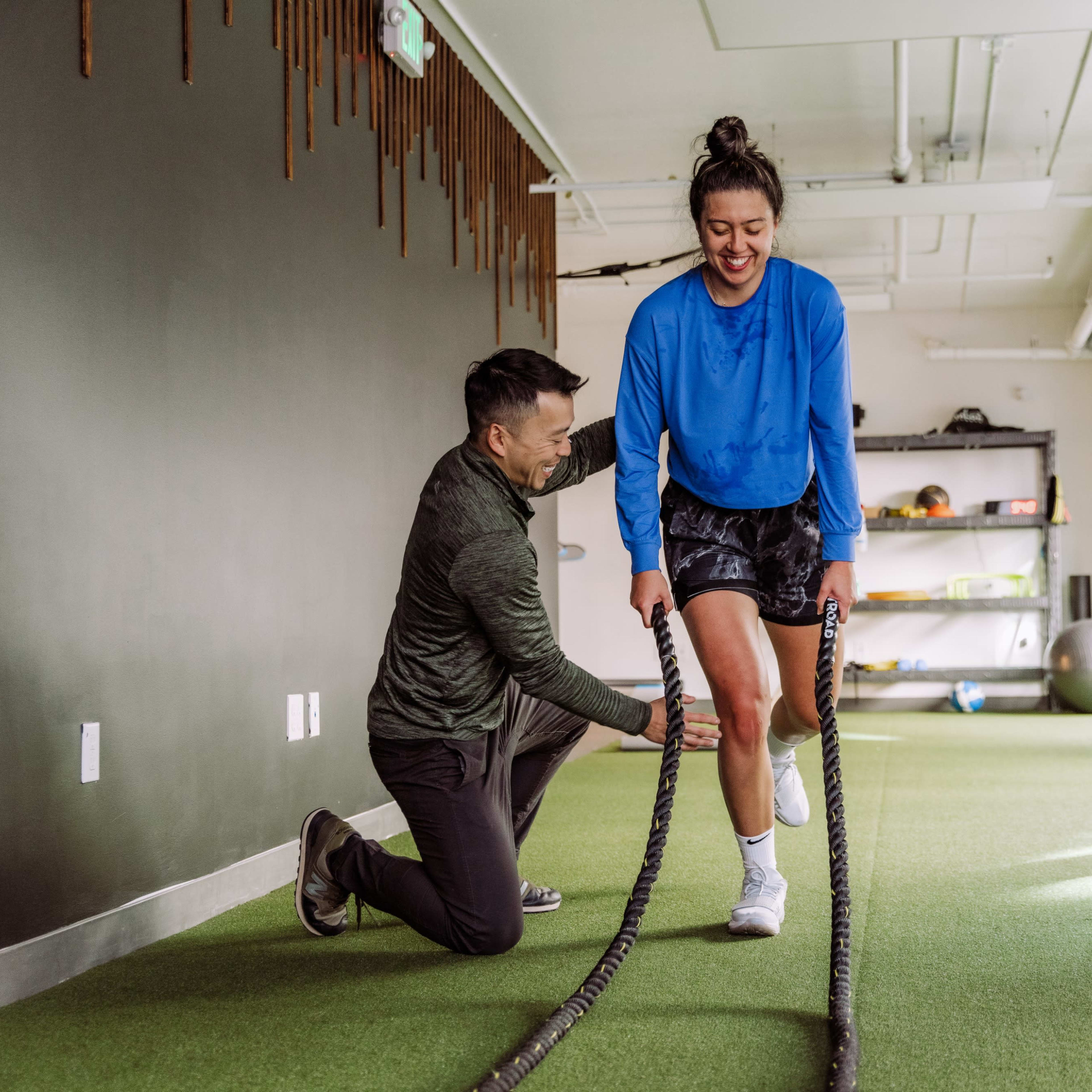 A trainer assists a woman in a workout using battle ropes in a gym setting.