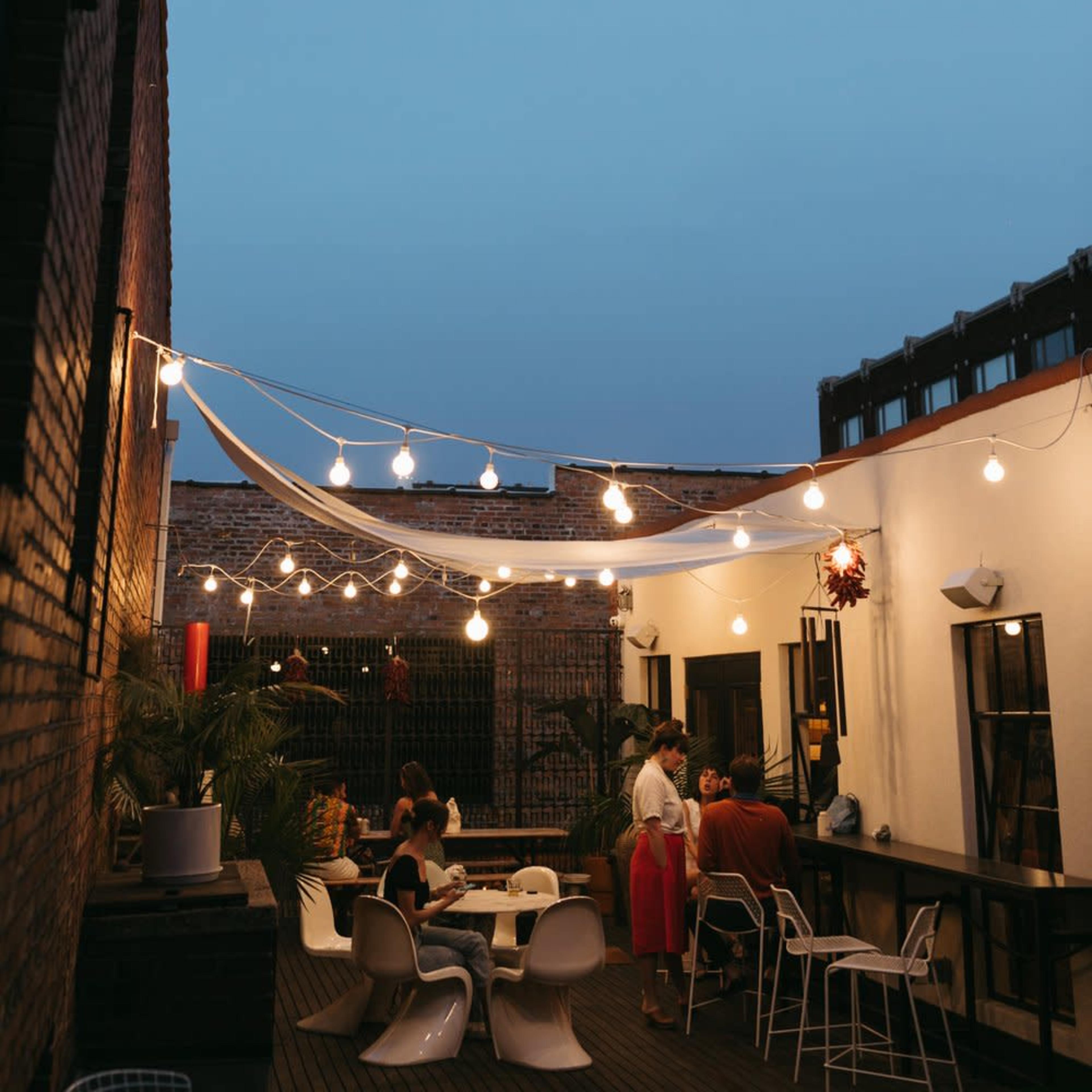 A rooftop terrace is illuminated by string lights, with several people gathered around tables and enjoying the evening.