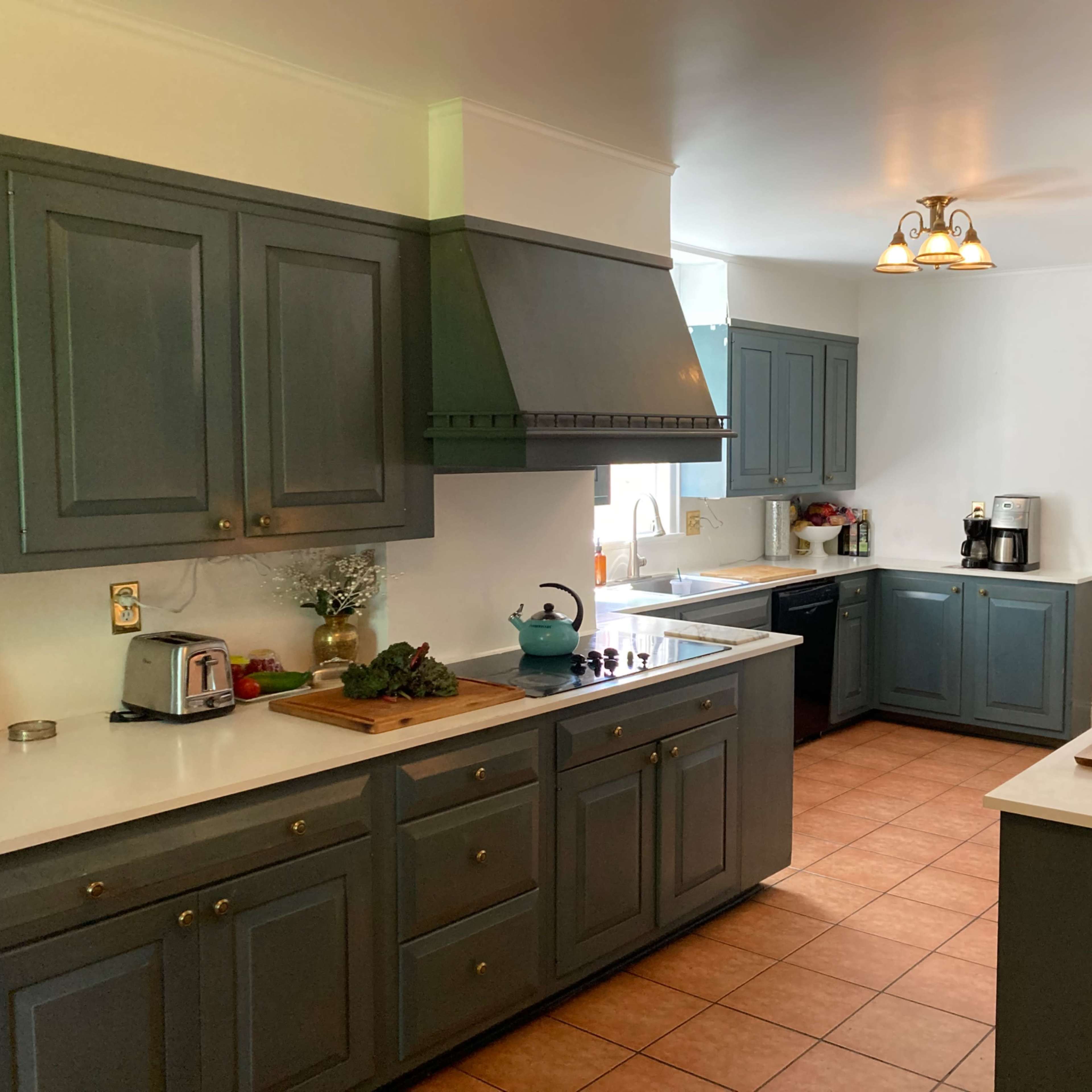 A kitchen with gray cabinets, a white countertop, and a tiled floor, featuring appliances and natural light from a window.
