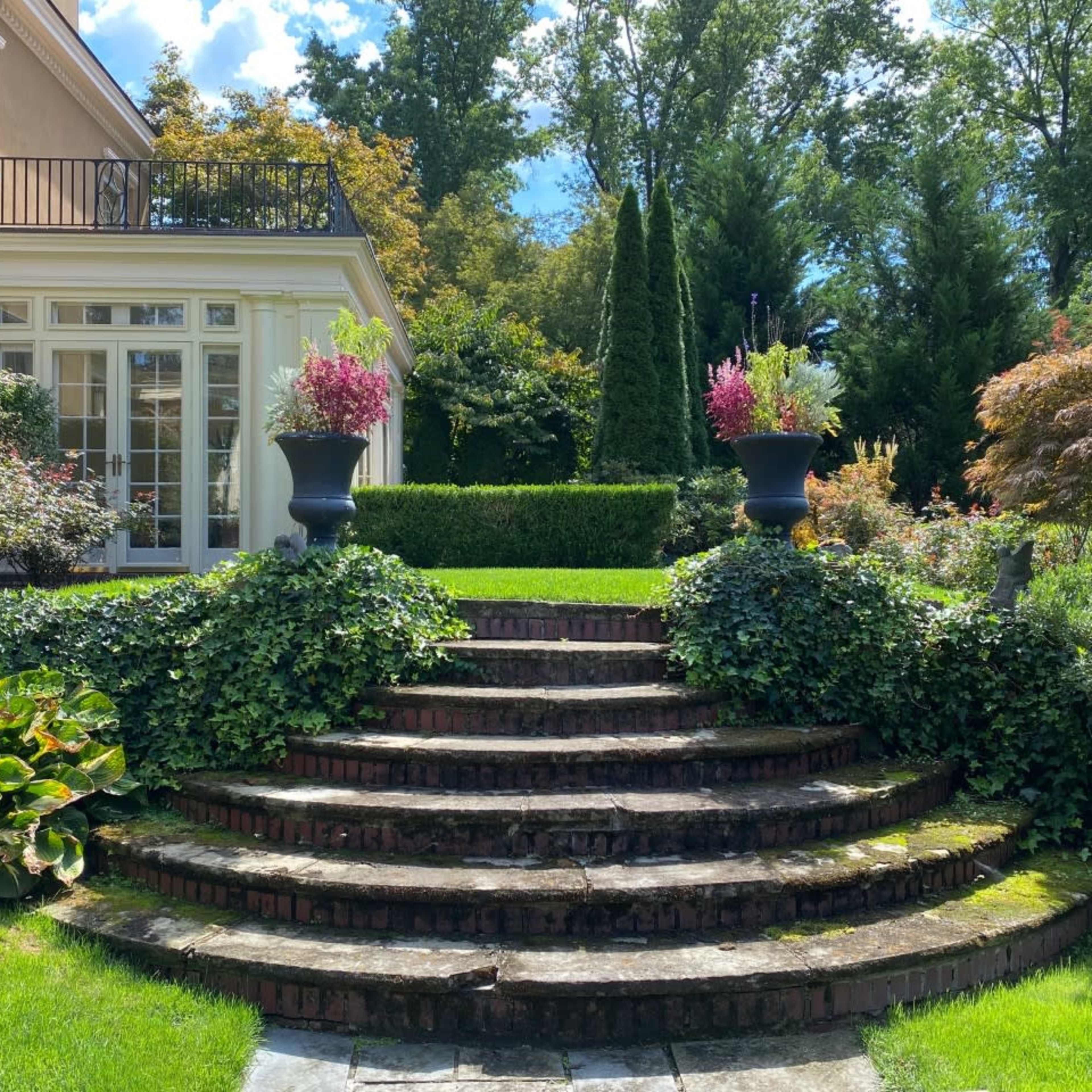 A landscaped garden with stone steps leading up to a house, flanked by ornamental plants and flowering urns on either side.