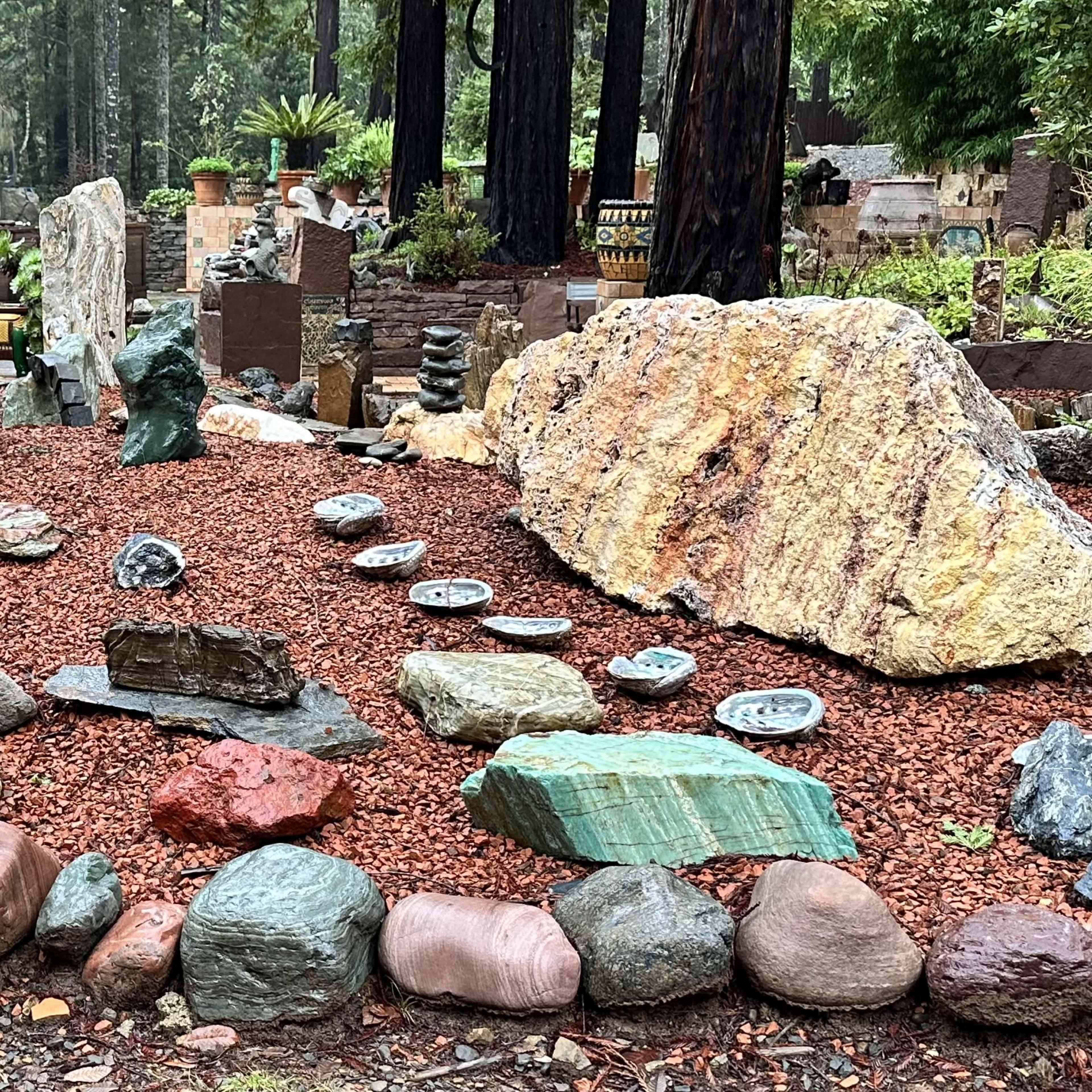 A landscaped area featuring various sizes and types of rocks, boulders, and decorative items arranged on a bed of red mulch.