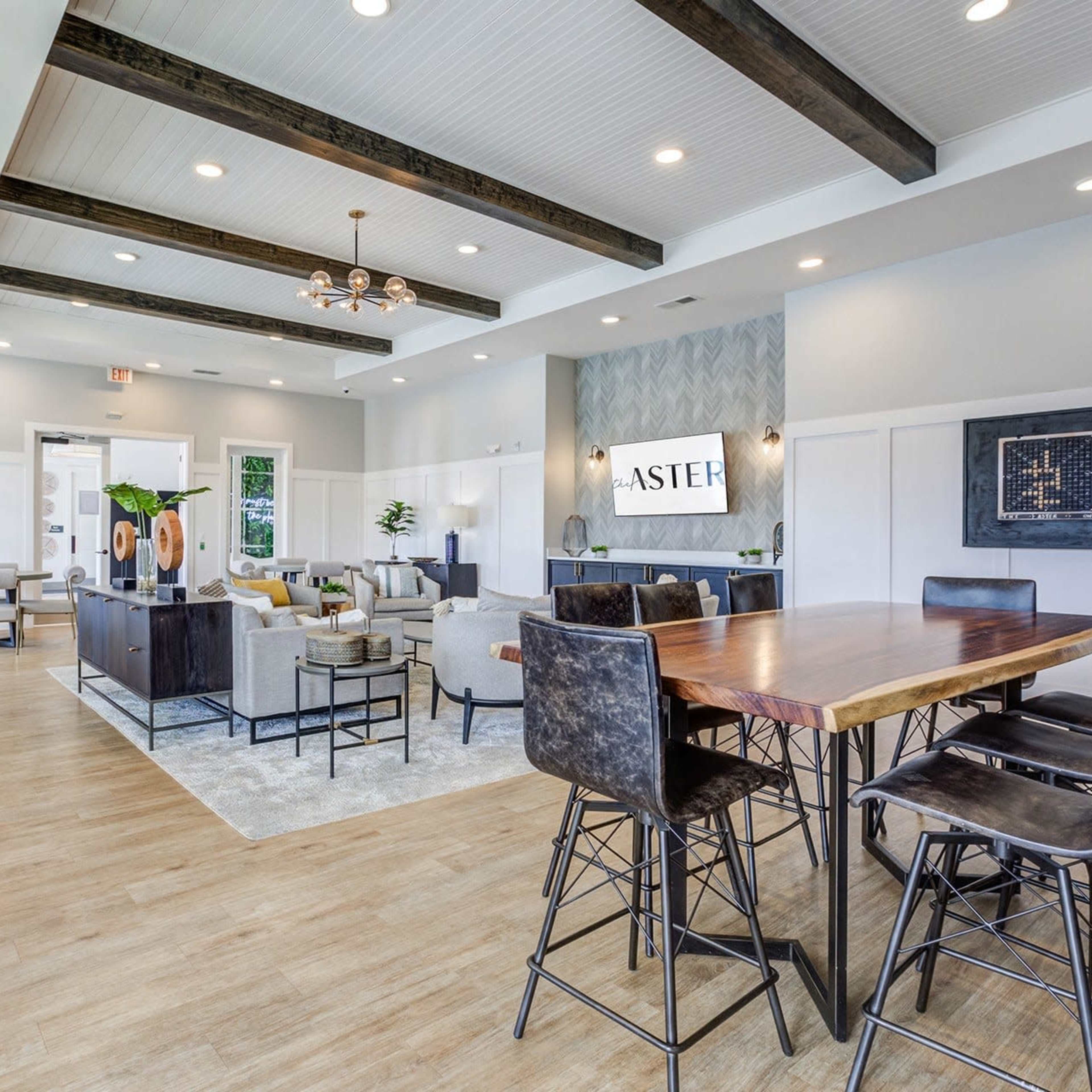 An interior lounge area with modern furniture, wooden beams on the ceiling, and a large communal table surrounded by high chairs.