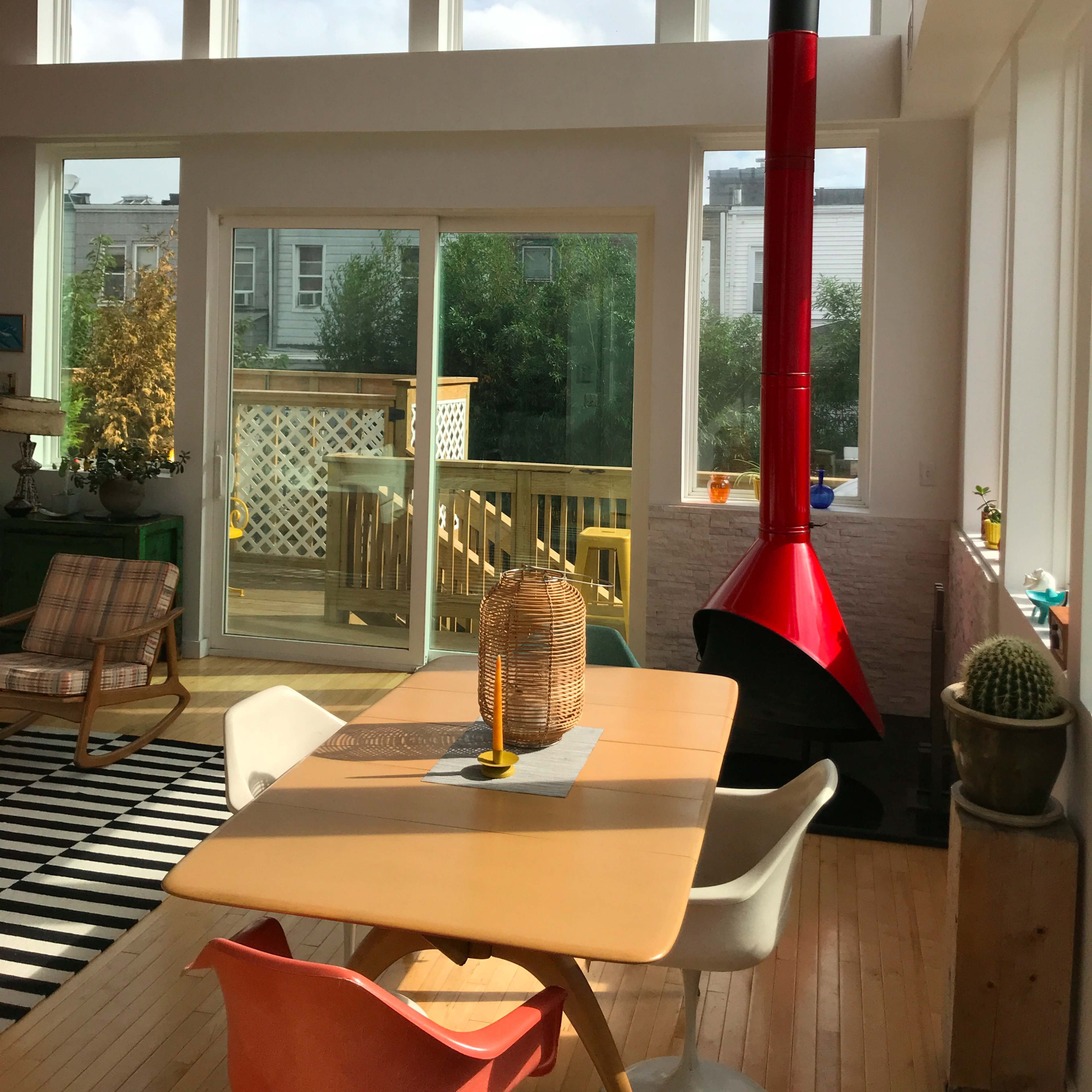 A bright, modern living space featuring large windows, a red chimney, and a wooden dining table surrounded by colorful chairs.
