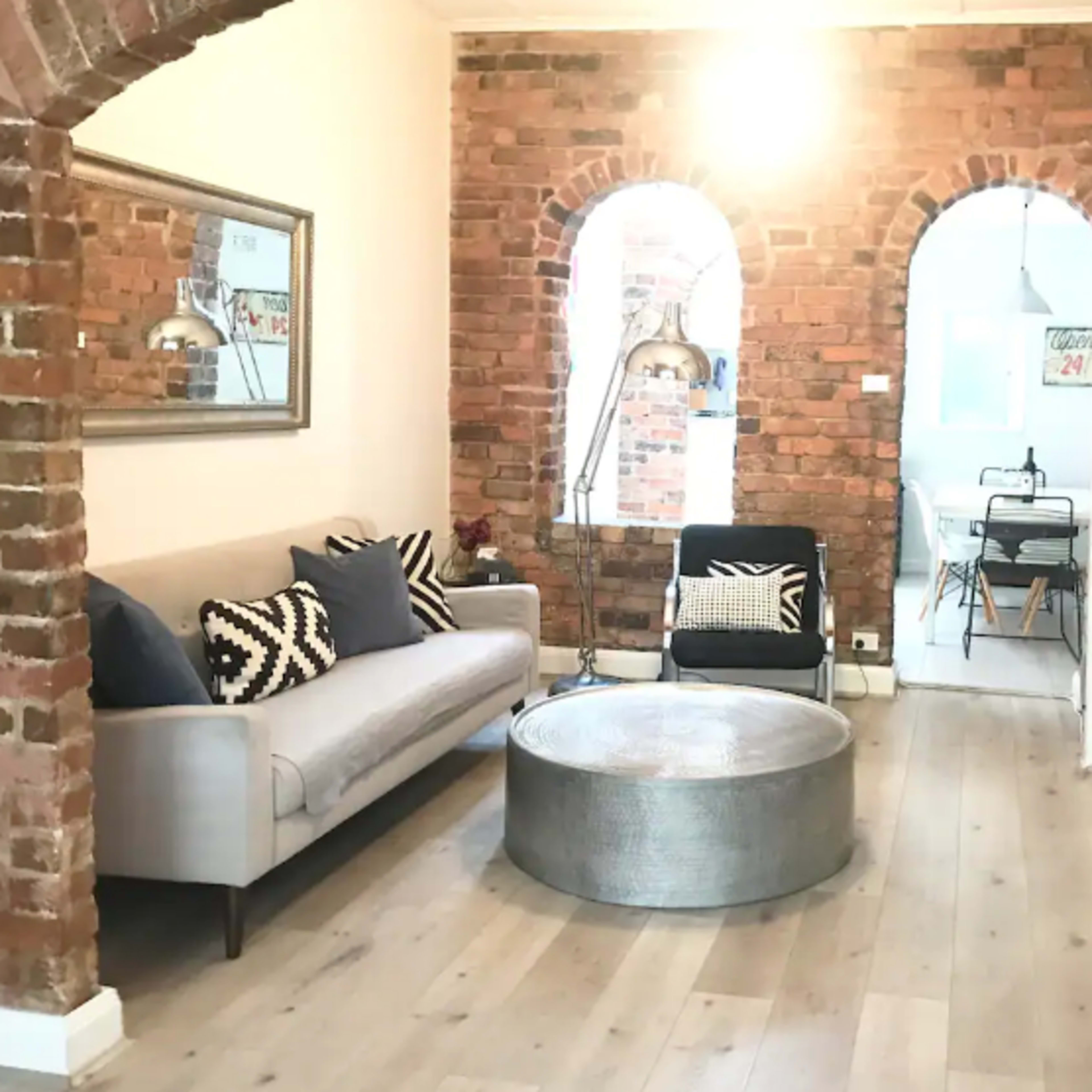 A living room with exposed brick walls, a gray sofa, and a round metallic coffee table, leading into a dining area visible in the background.