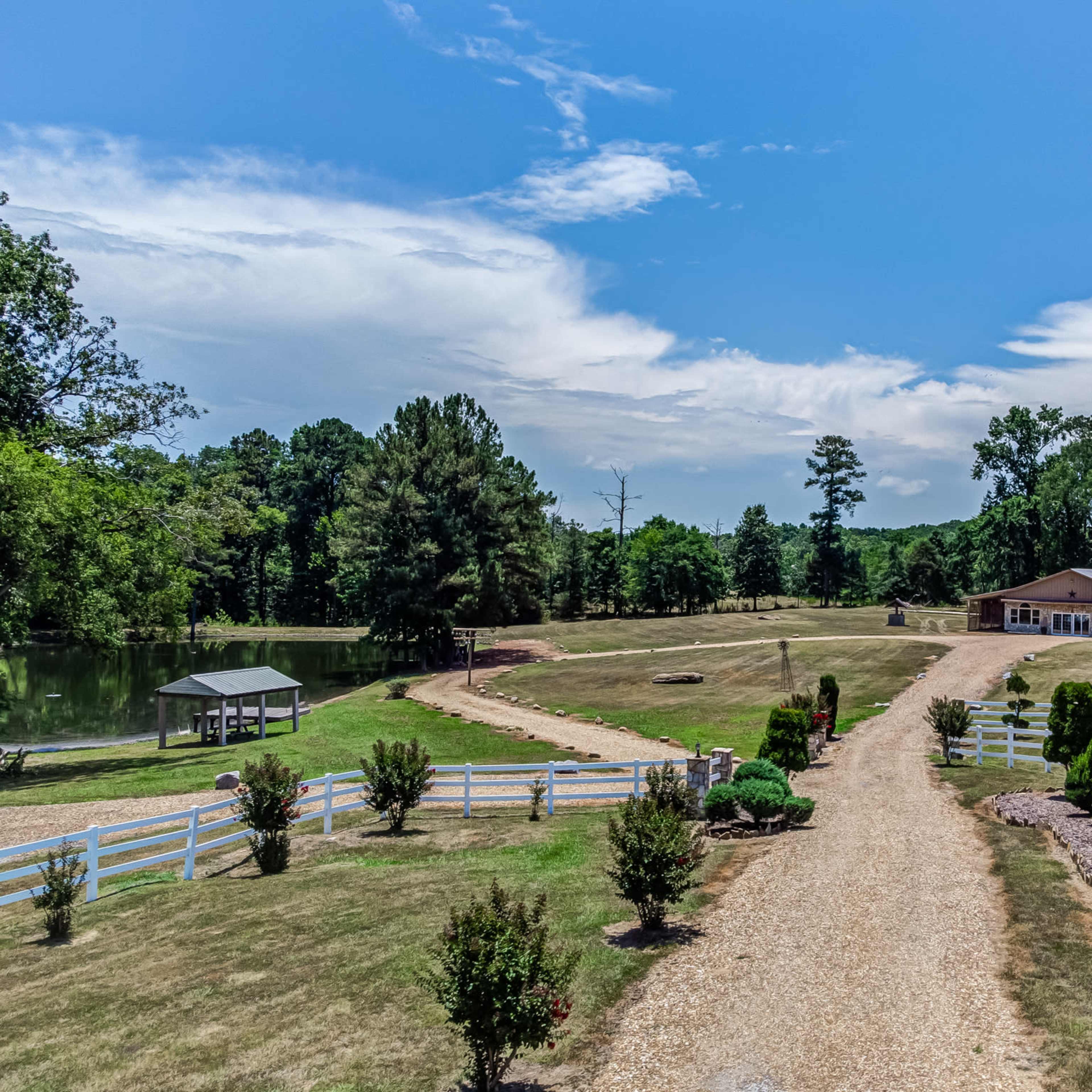 A spacious landscape with a gravel driveway leading to a large building, surrounded by manicured greenery and a pond on the left.