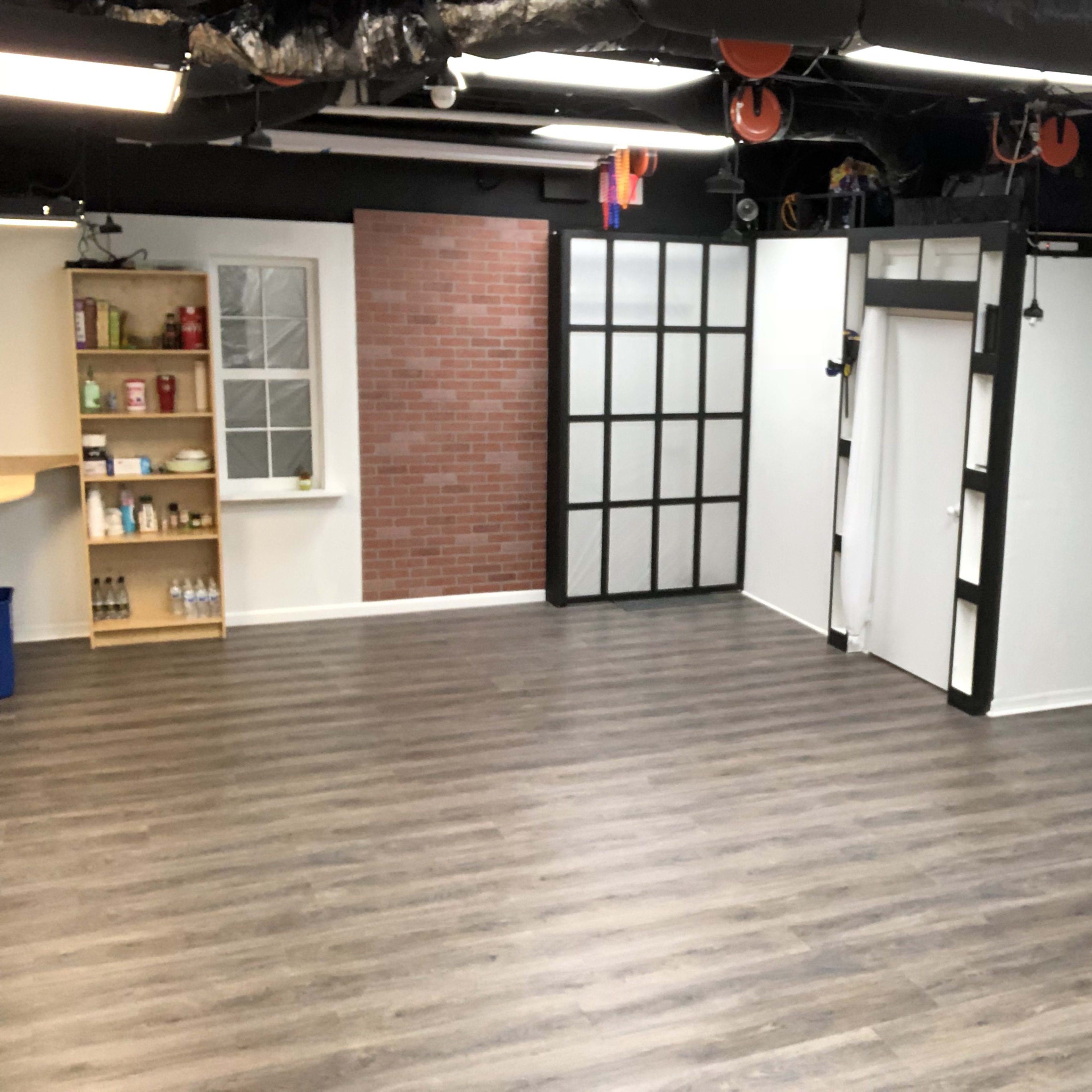 An empty room with a wooden floor, featuring exposed walls with a brick section, glass-paneled doors, and a shelf stocked with items against one wall.