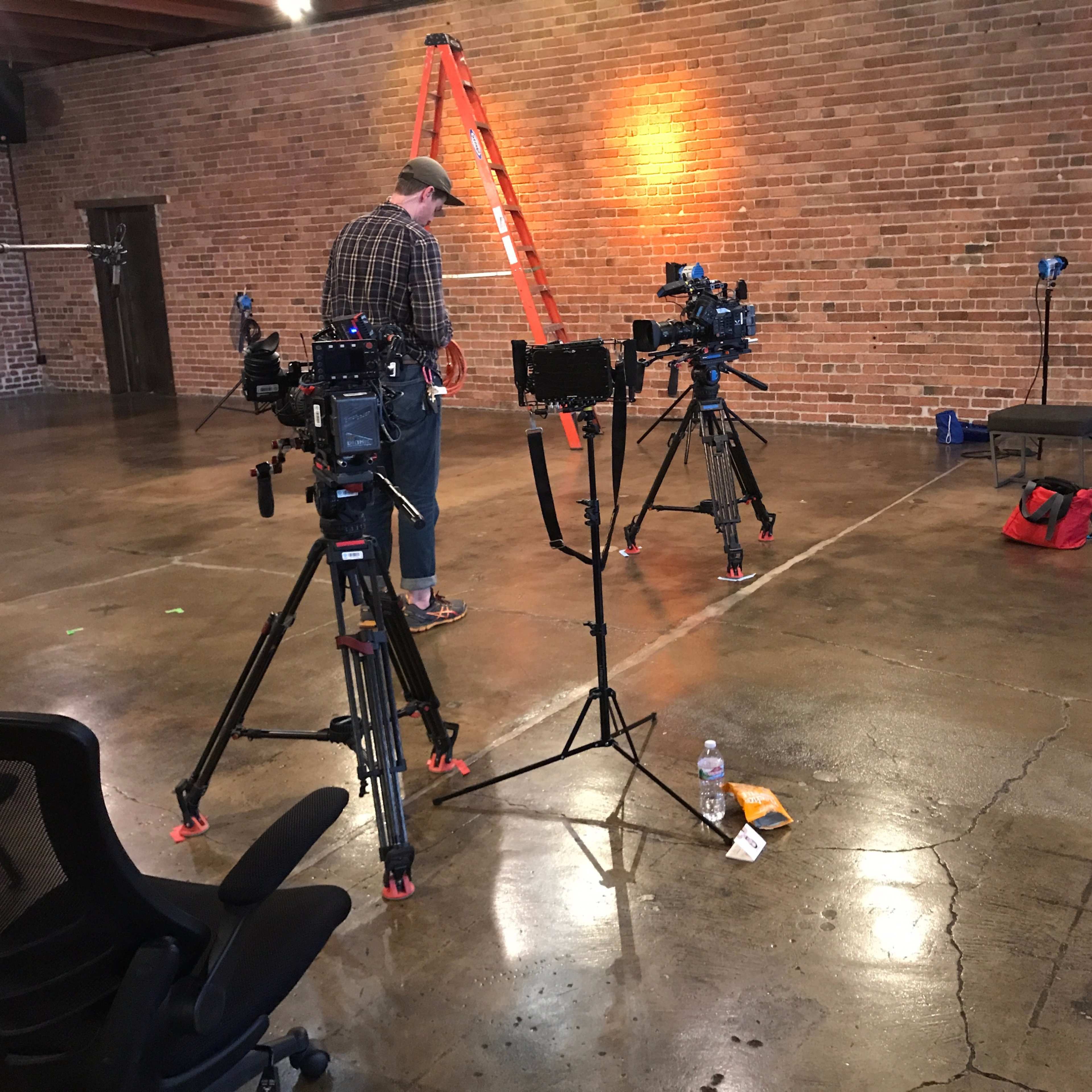 A man sets up camera equipment in a spacious, empty industrial-style room with brick walls and a polished concrete floor.