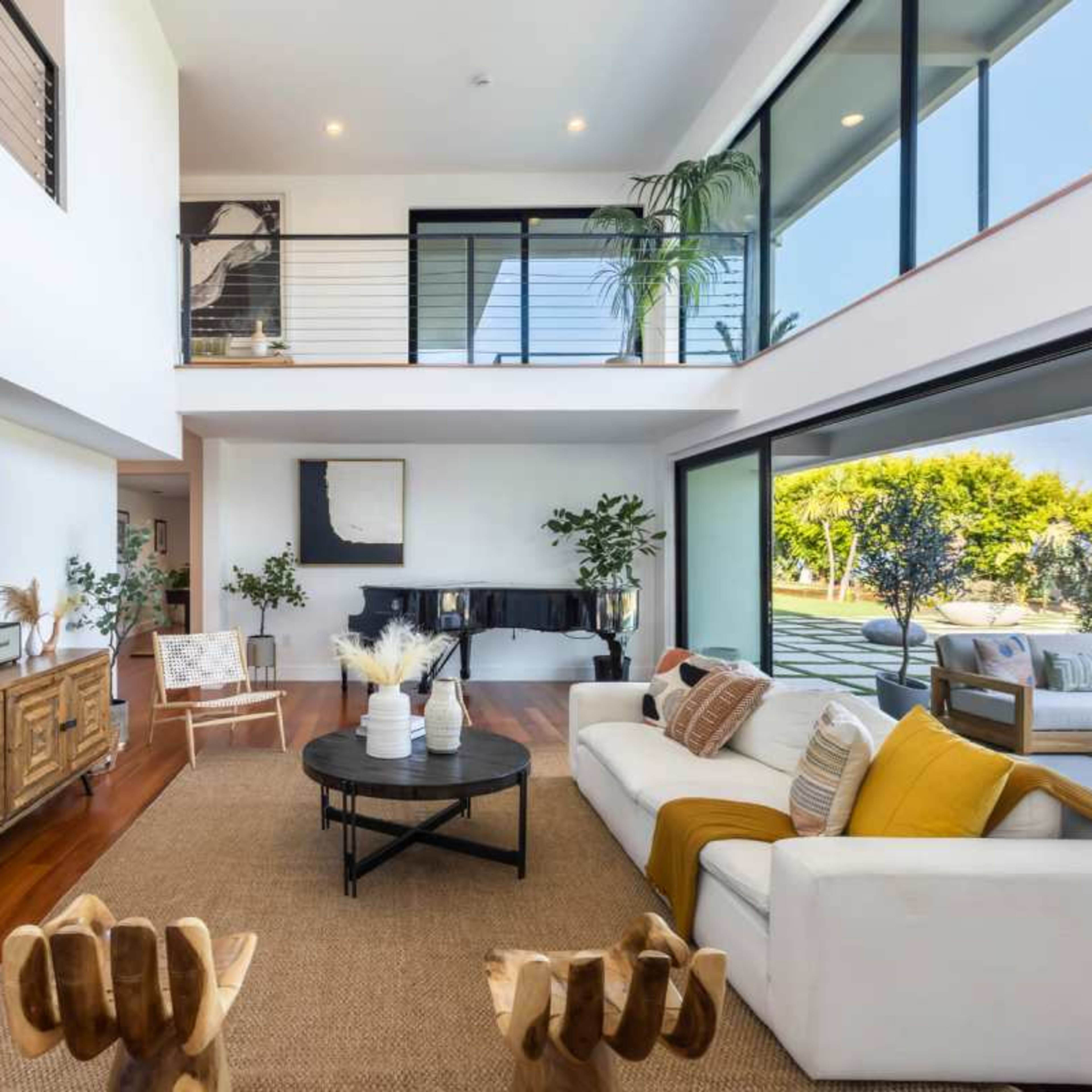 A modern living room with large windows, a wooden coffee table, a white sofa with yellow and beige pillows, and indoor plants.
