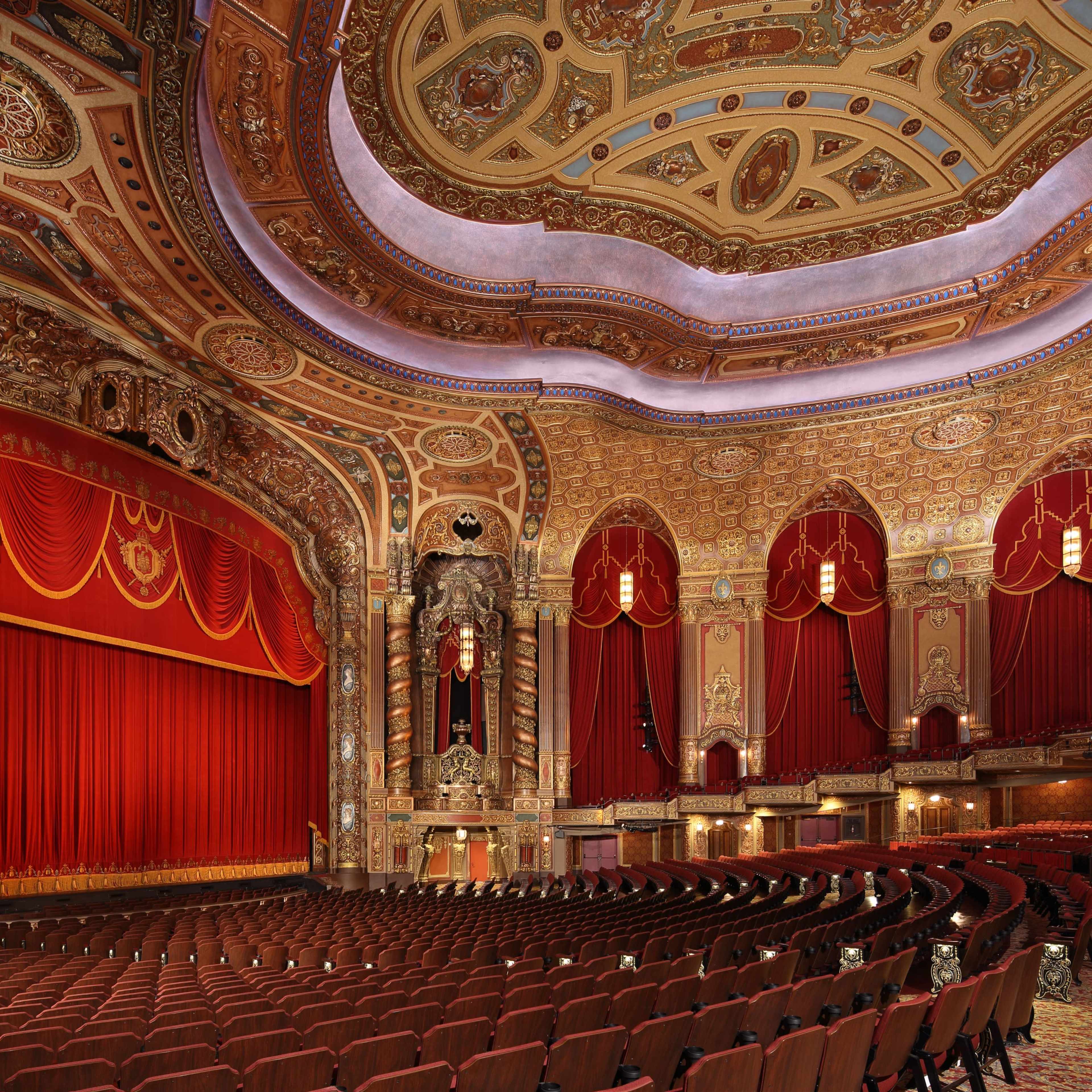 An ornate theater interior with a red curtain, intricate ceiling designs, and rows of maroon seats.
