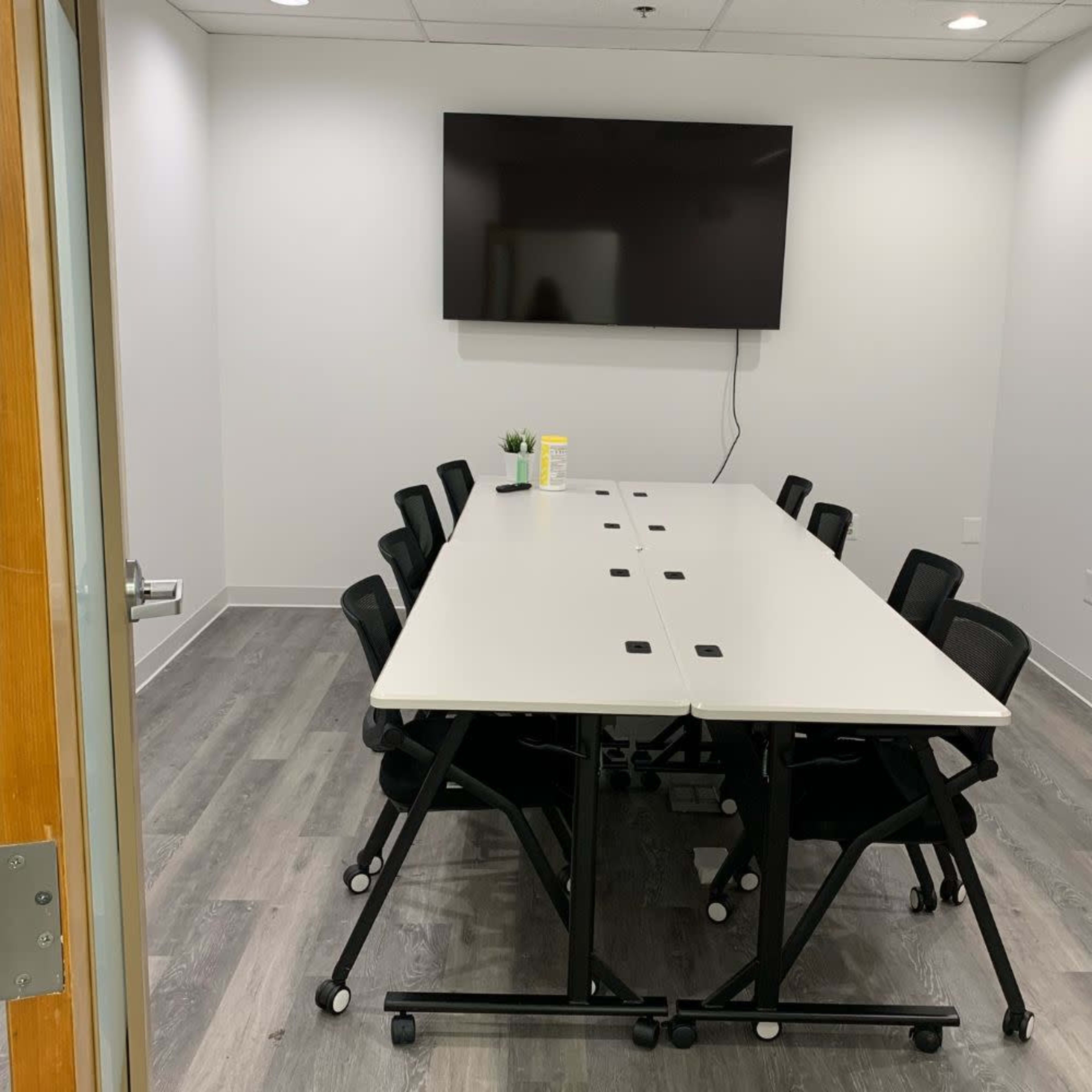 A conference room with a long white table surrounded by black chairs, featuring a large wall-mounted television.