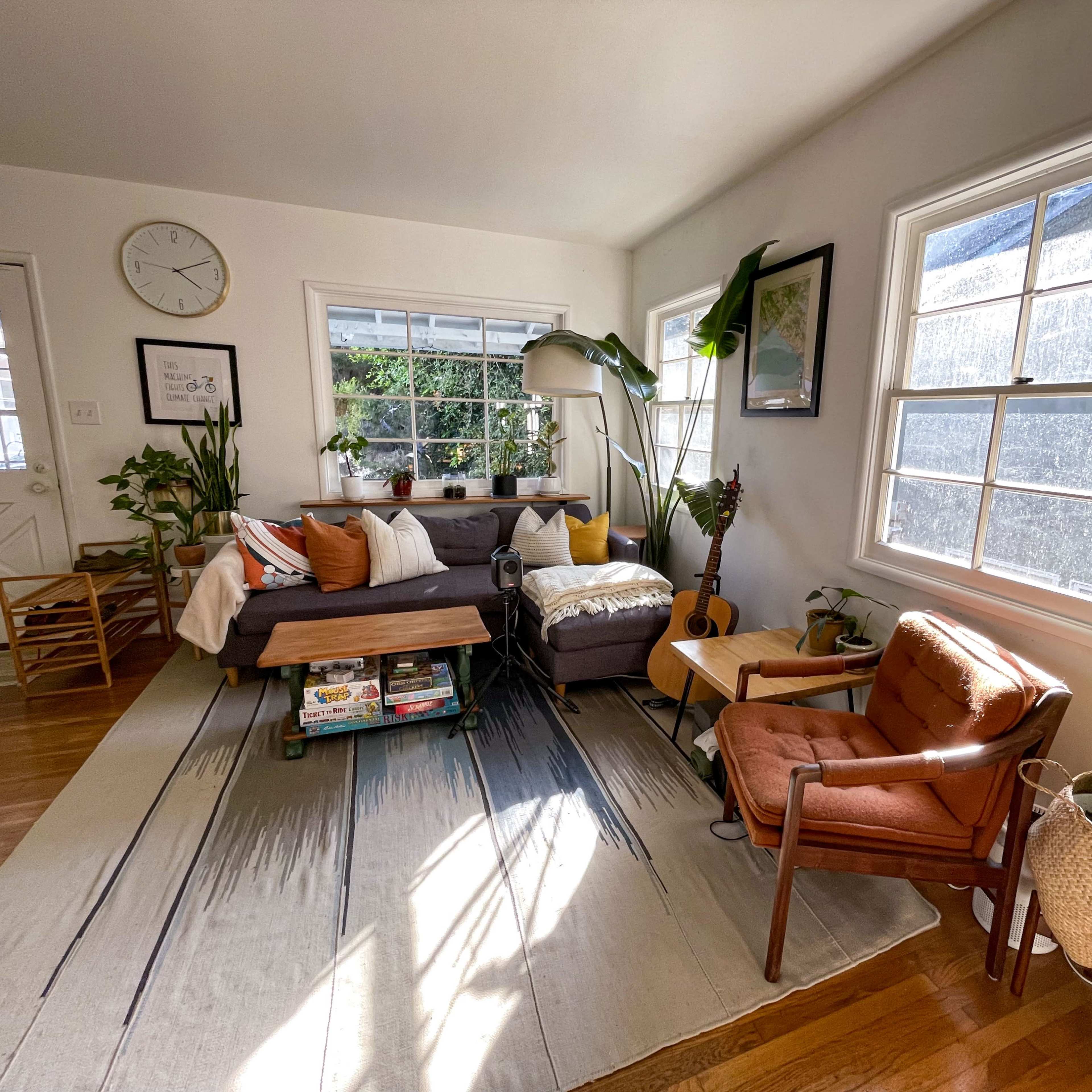 A cozy living room features a mix of plants, a sofa with colorful cushions, a guitar, and large windows allowing natural light.