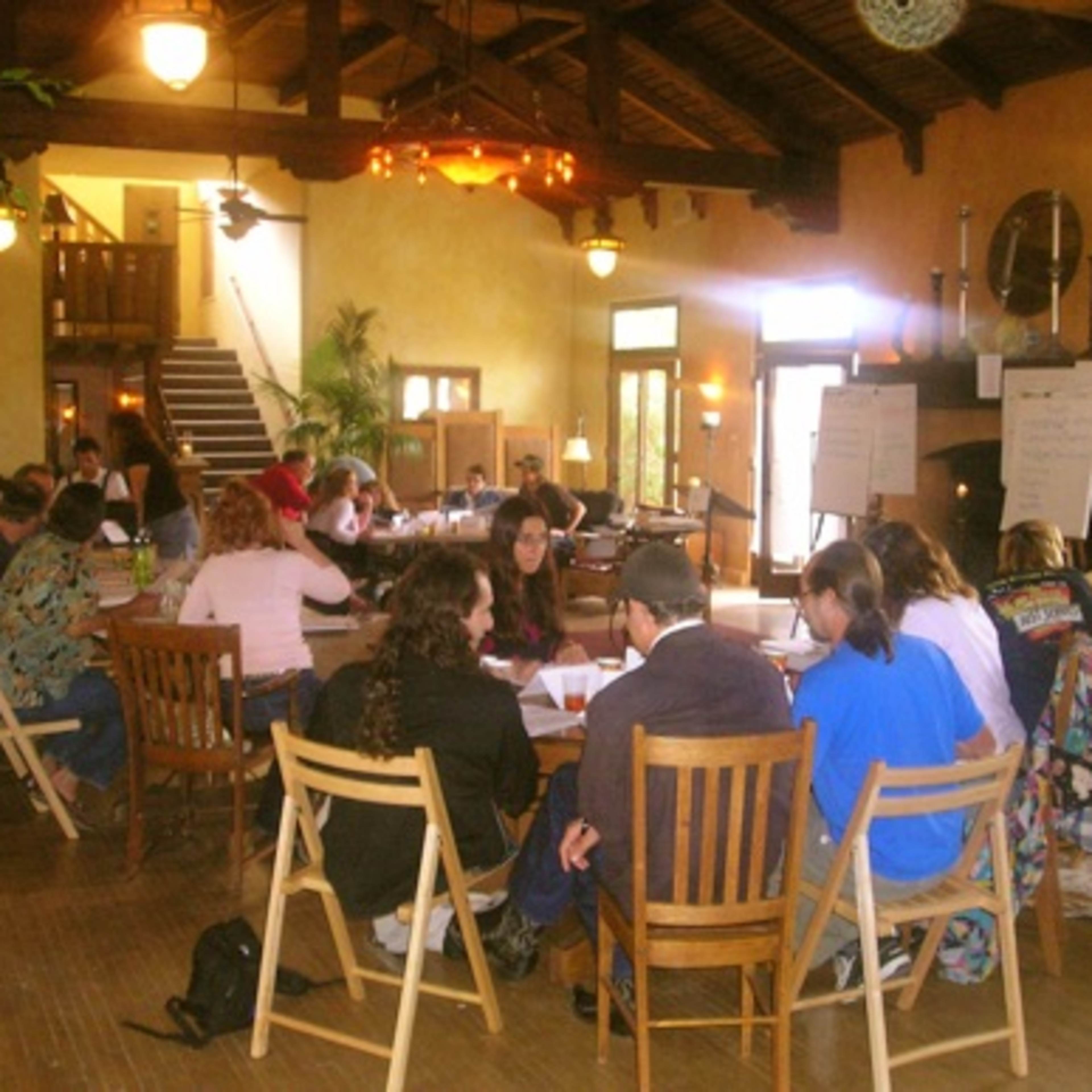A group of people is seated around tables in a spacious room with high ceilings, participating in a meeting or workshop.
