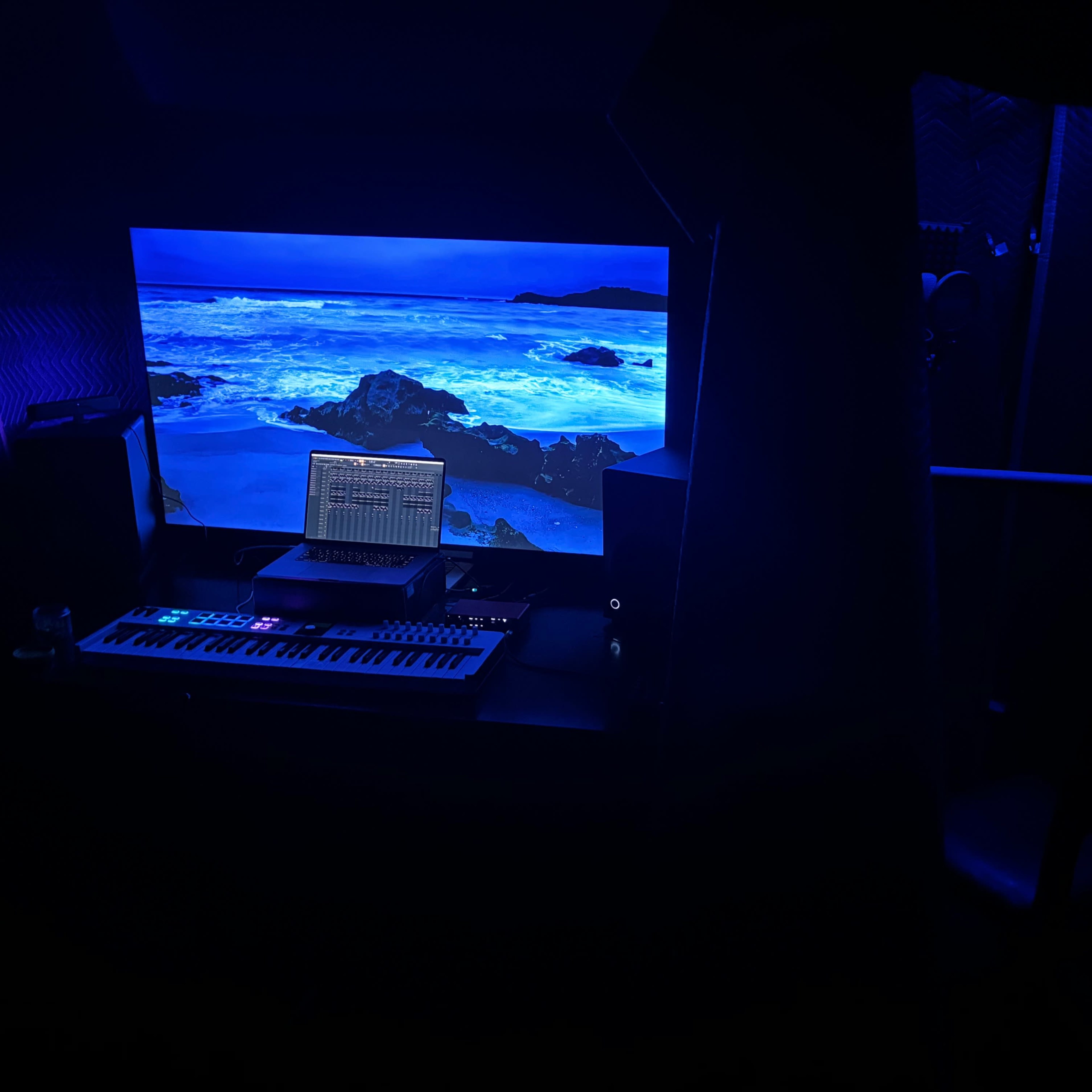 A dimly lit music production setup featuring a keyboard, a laptop displaying audio software, and a large screen with a coastal landscape.
