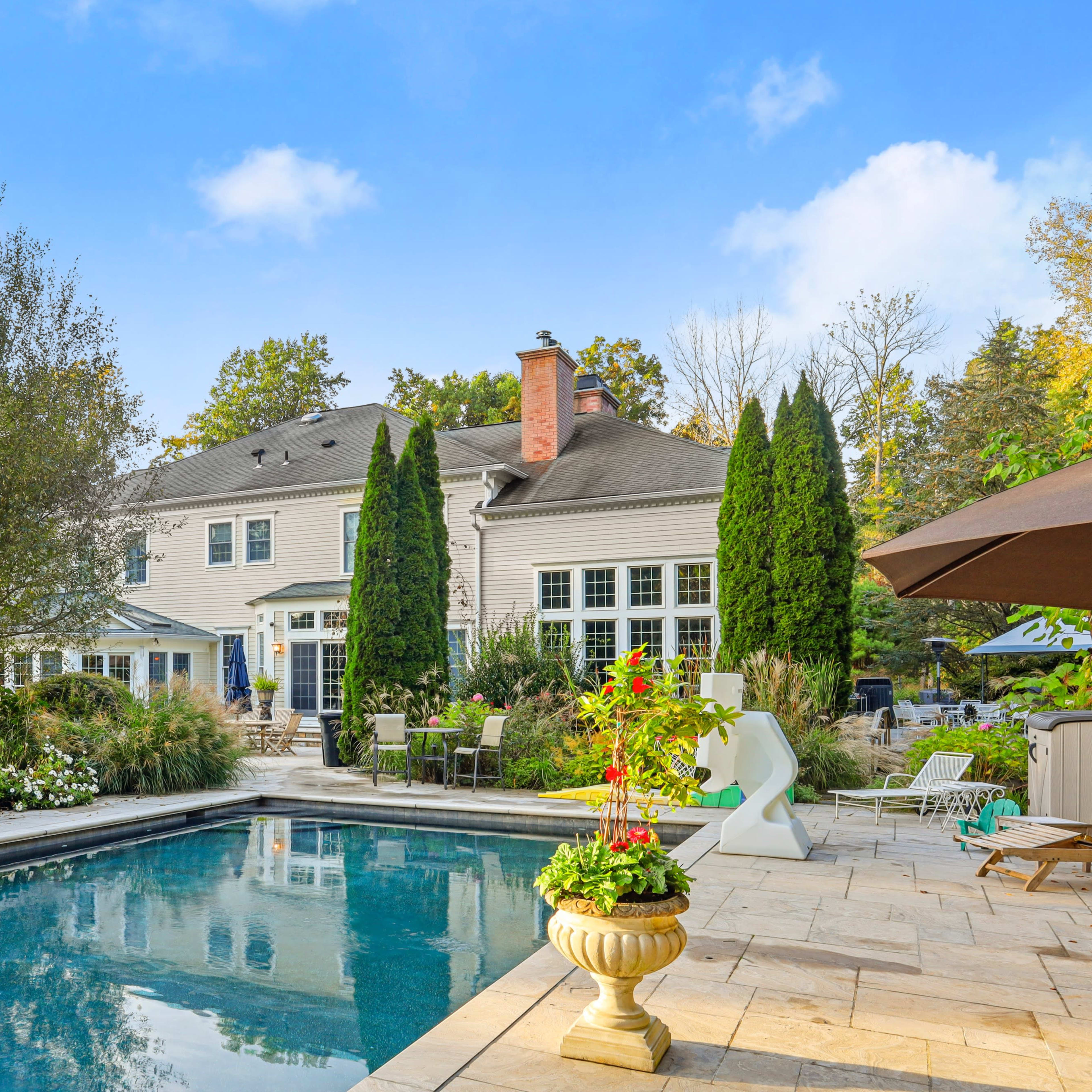 A backyard with a swimming pool in the foreground, a large house in the background, and landscaped gardens with trees and seating areas.