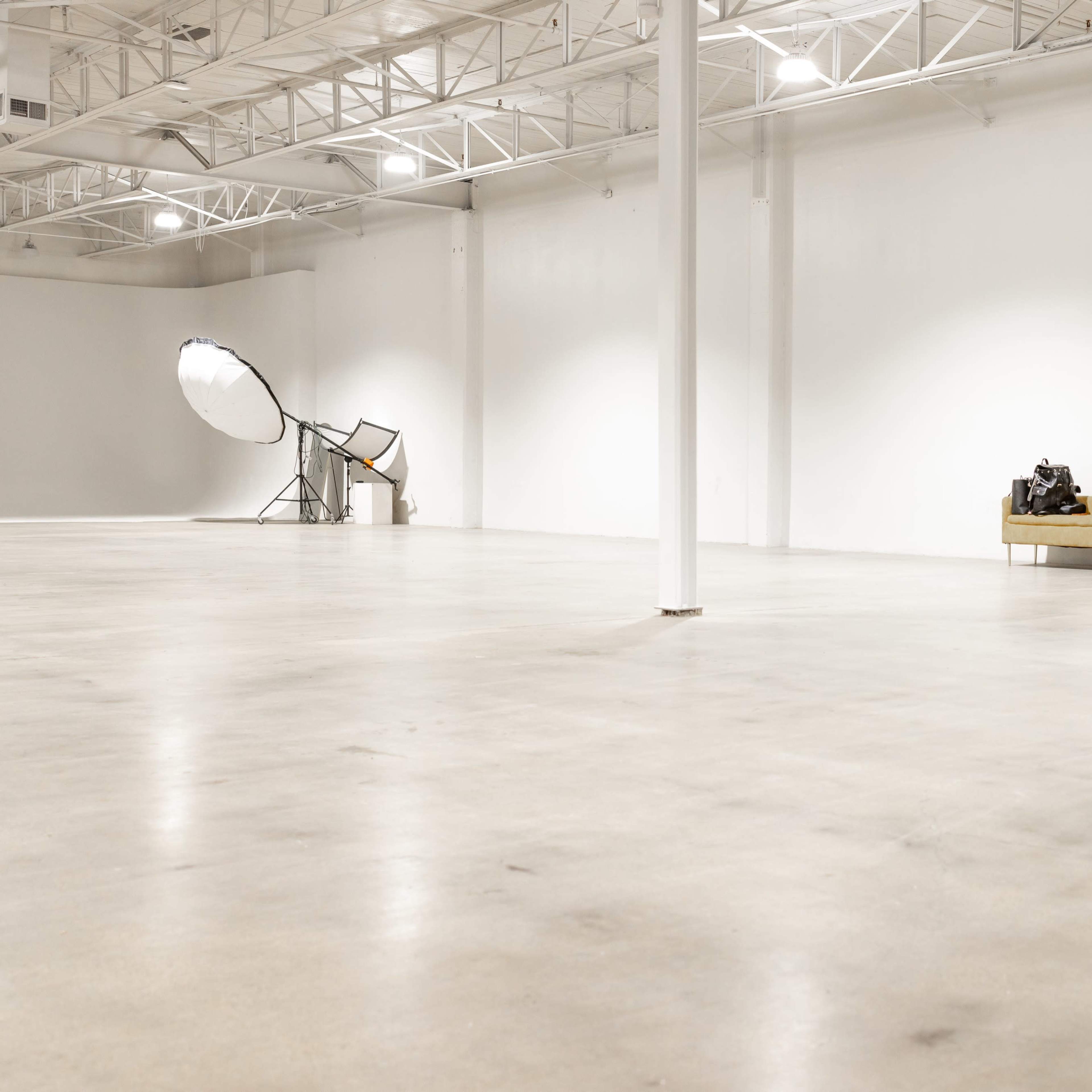An empty industrial space with a polished concrete floor, a sofa against one wall, and large lighting equipment in the background.