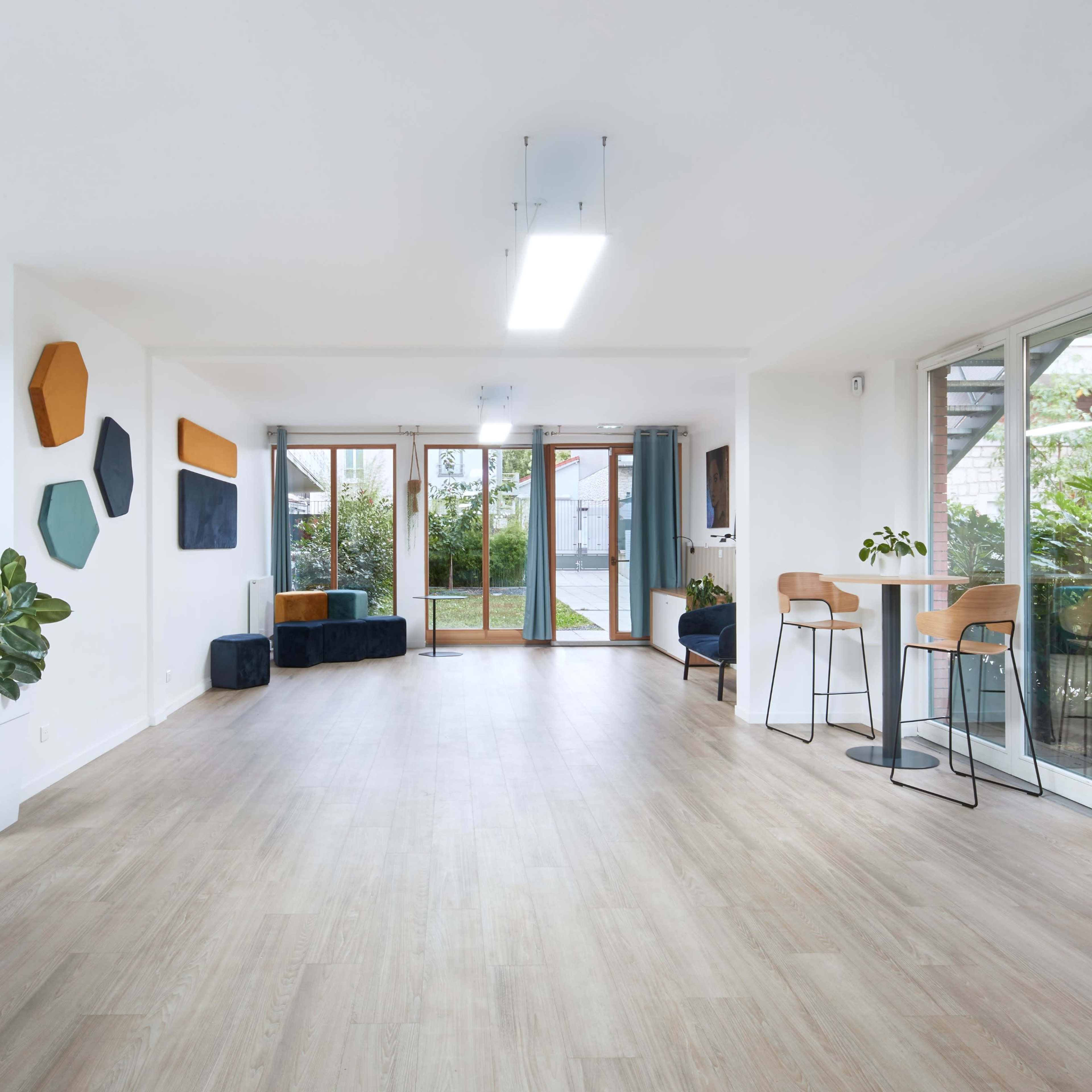 A spacious, well-lit interior features wooden flooring, modern seating arrangements, and large glass doors leading to an outdoor area.