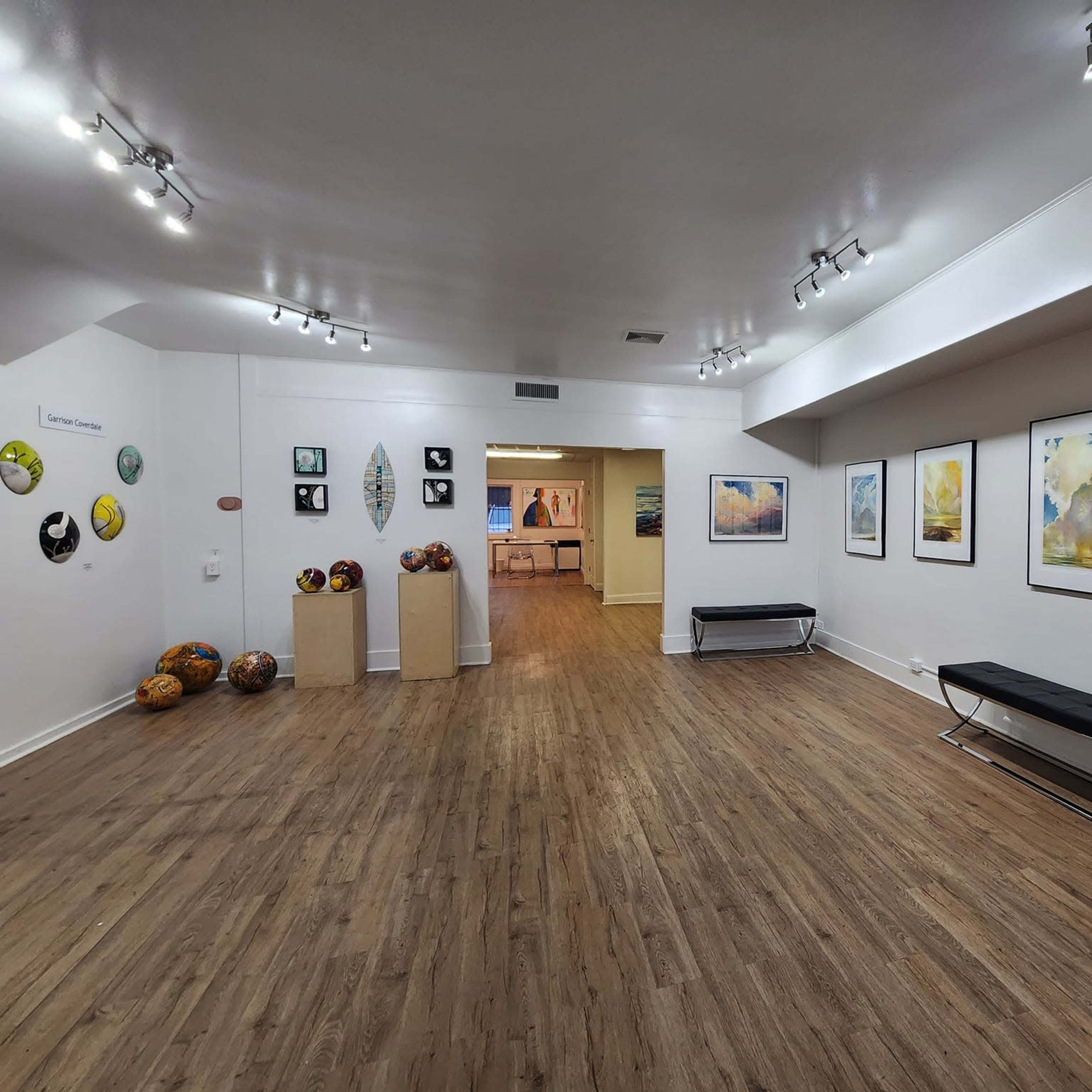 An art gallery featuring several paintings on white walls, a wooden floor, and a few sculptures displayed on pedestals.