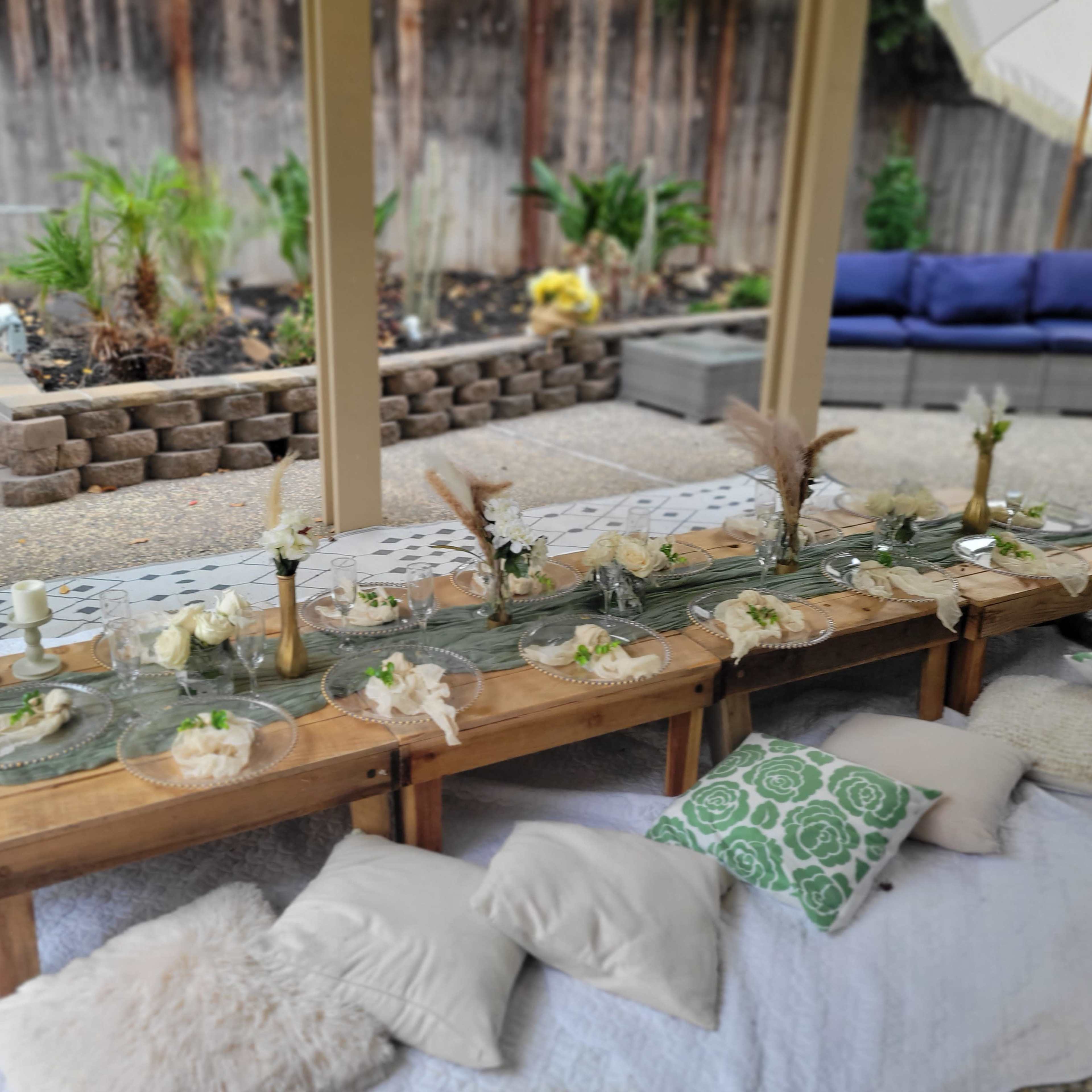 A long outdoor dining table set with glass plates, white napkins, and decorative accents, surrounded by cushions and plants.