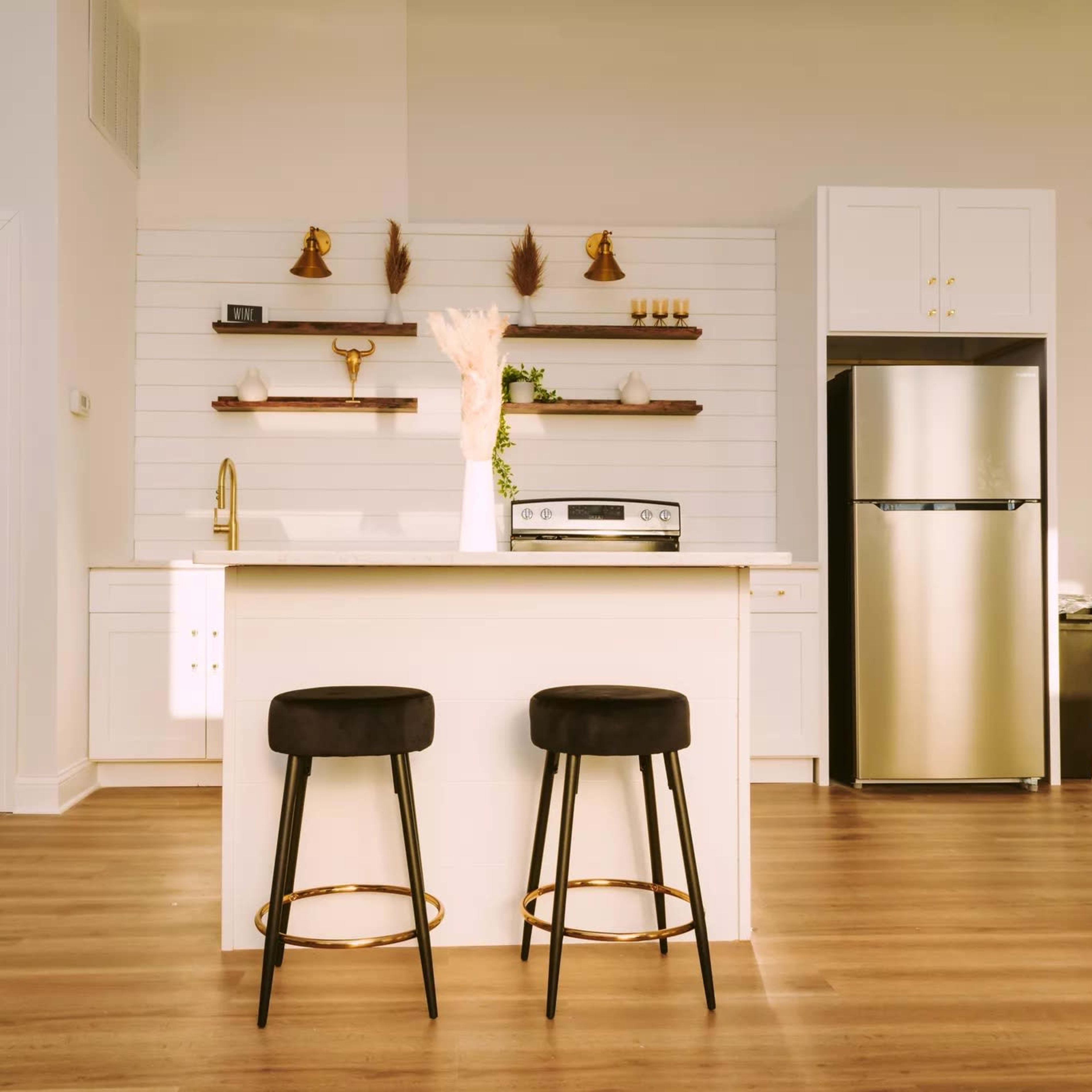 A modern kitchen features a central island with two black stools, minimalist shelves with decor, and stainless steel appliances.