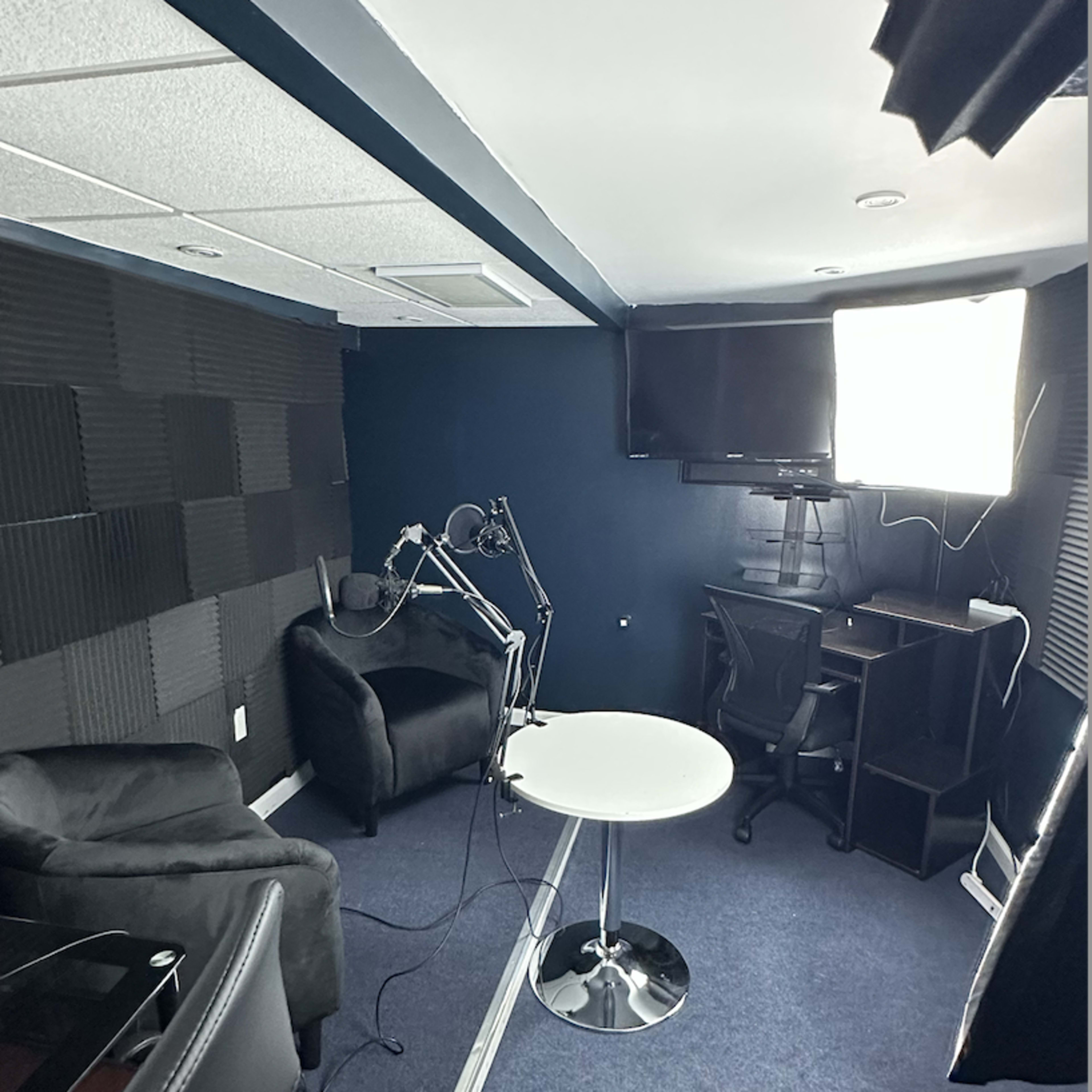 A small recording studio with black soundproofing panels, a round white table, two black chairs, and equipment including a monitor and a light.