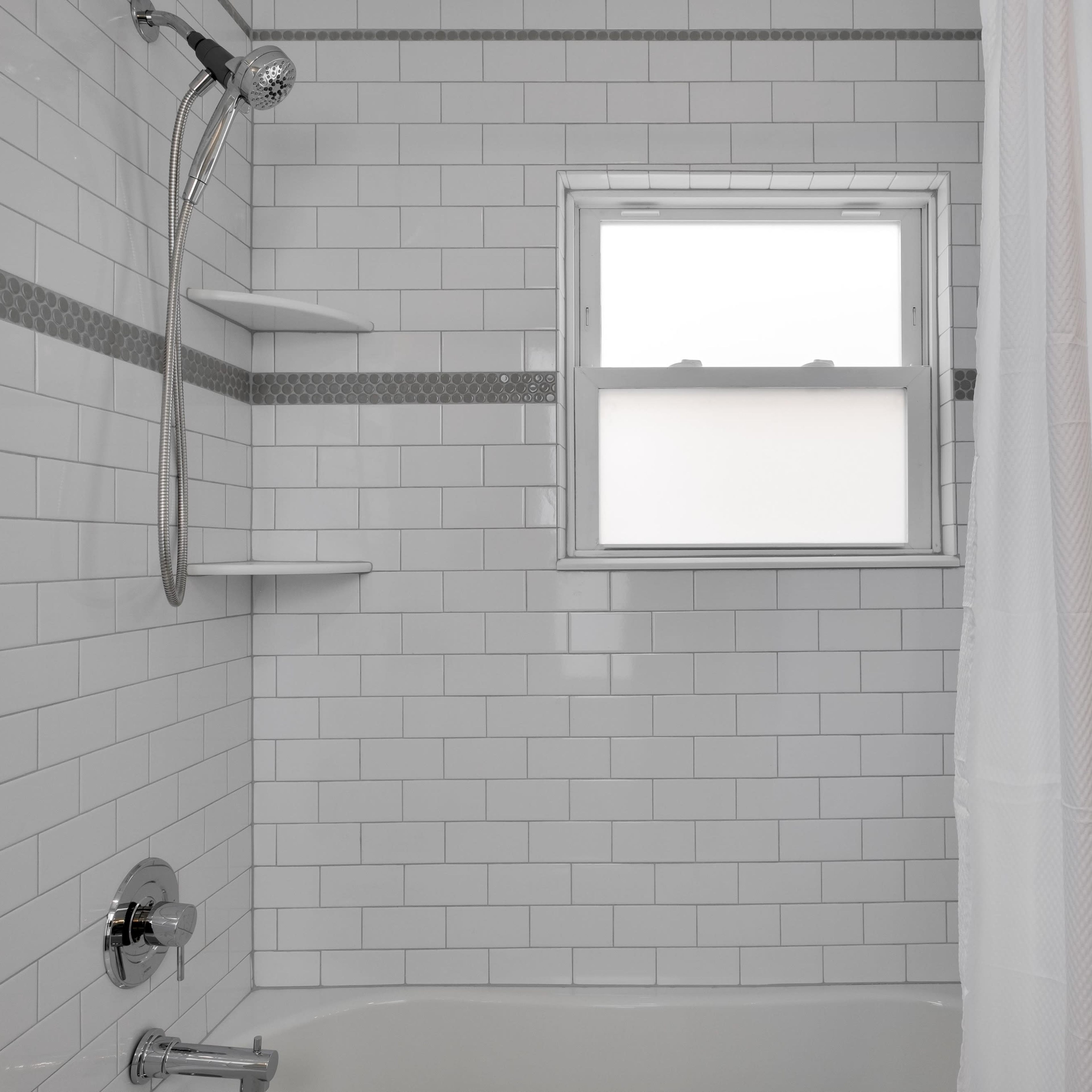 A clean, white tiled bathroom with a shower area, a bathtub, and a window.