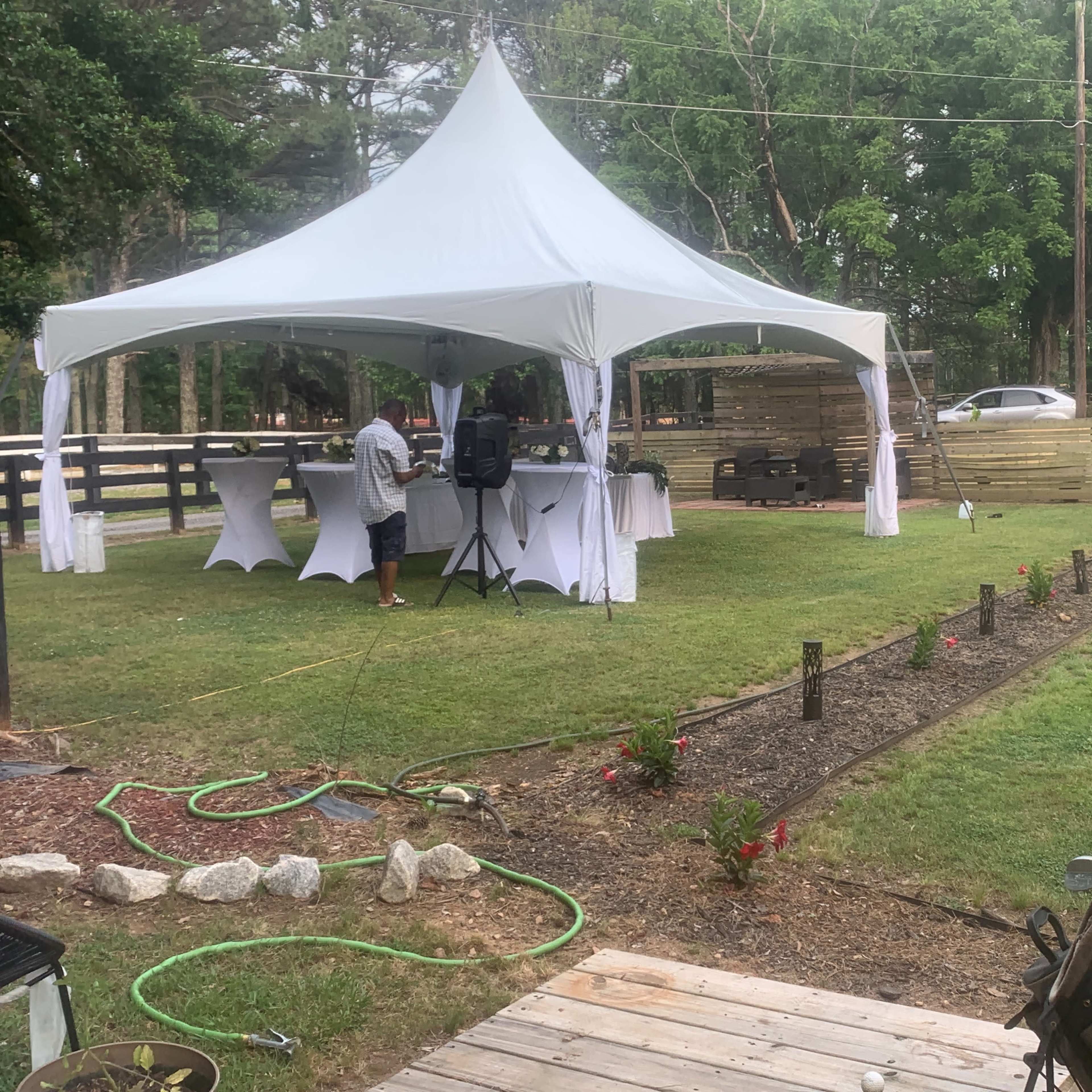 A white tent is set up on a grassy area with tables underneath, while a person prepares for an event in the background, and a pathway lined with flowers leads through the yard.