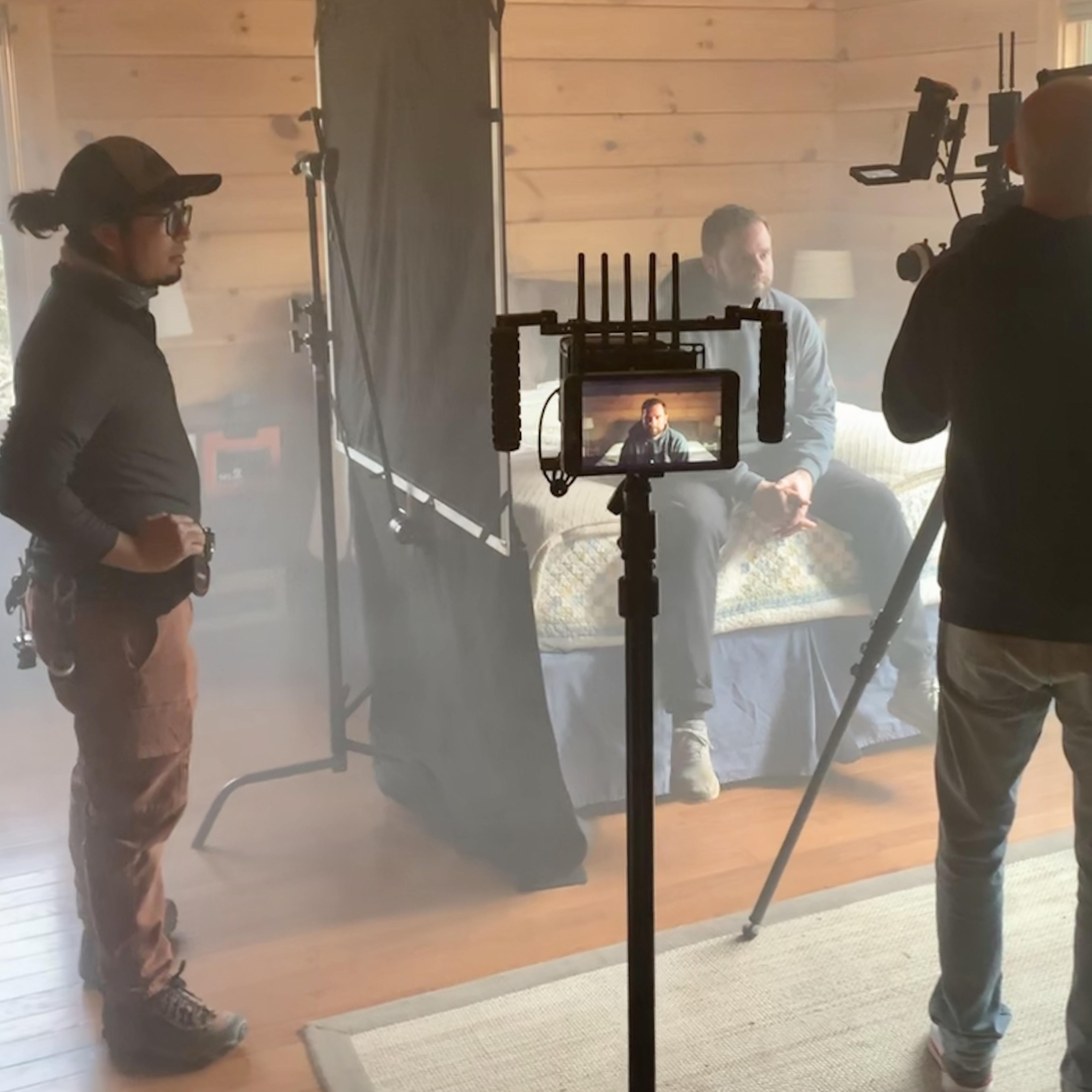A film crew sets up a shot in a wooden room with fog, focusing on a seated subject while using multiple cameras and lights.