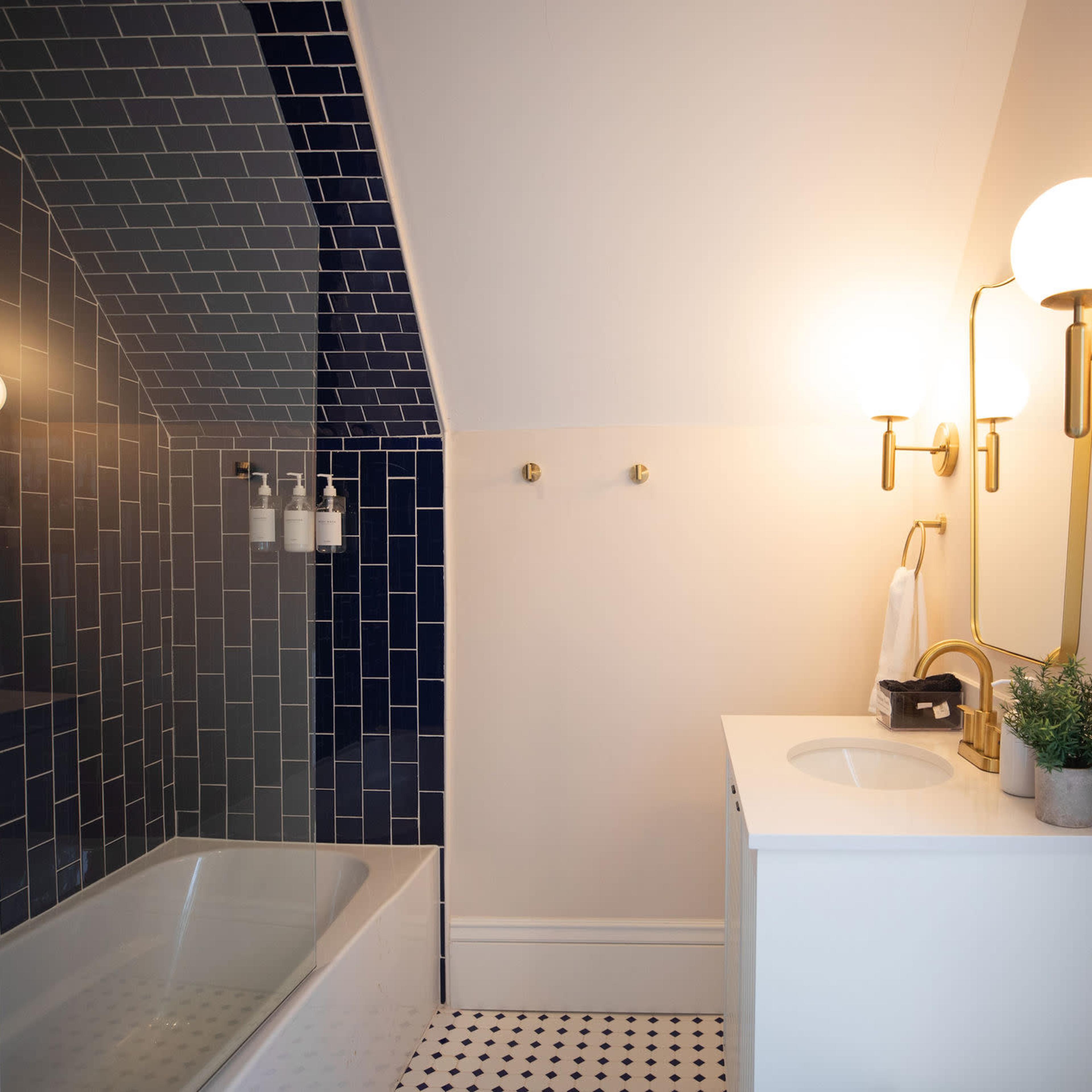 A modern bathroom featuring a bathtub with a blue tiled wall, a white sink with a gold faucet, and two wall-mounted light fixtures.