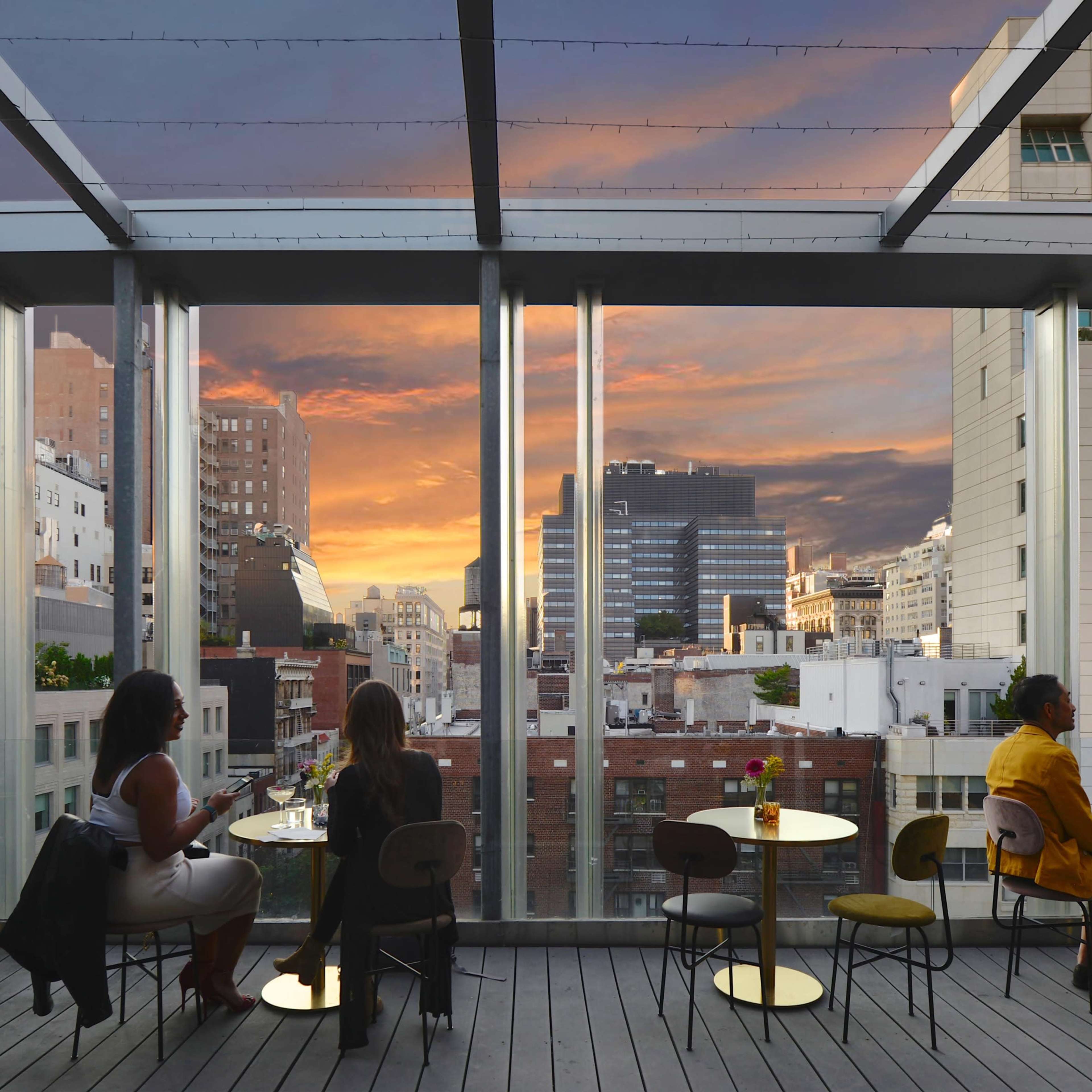 A rooftop terrace features patrons seated at tables, overlooking a city skyline during sunset.