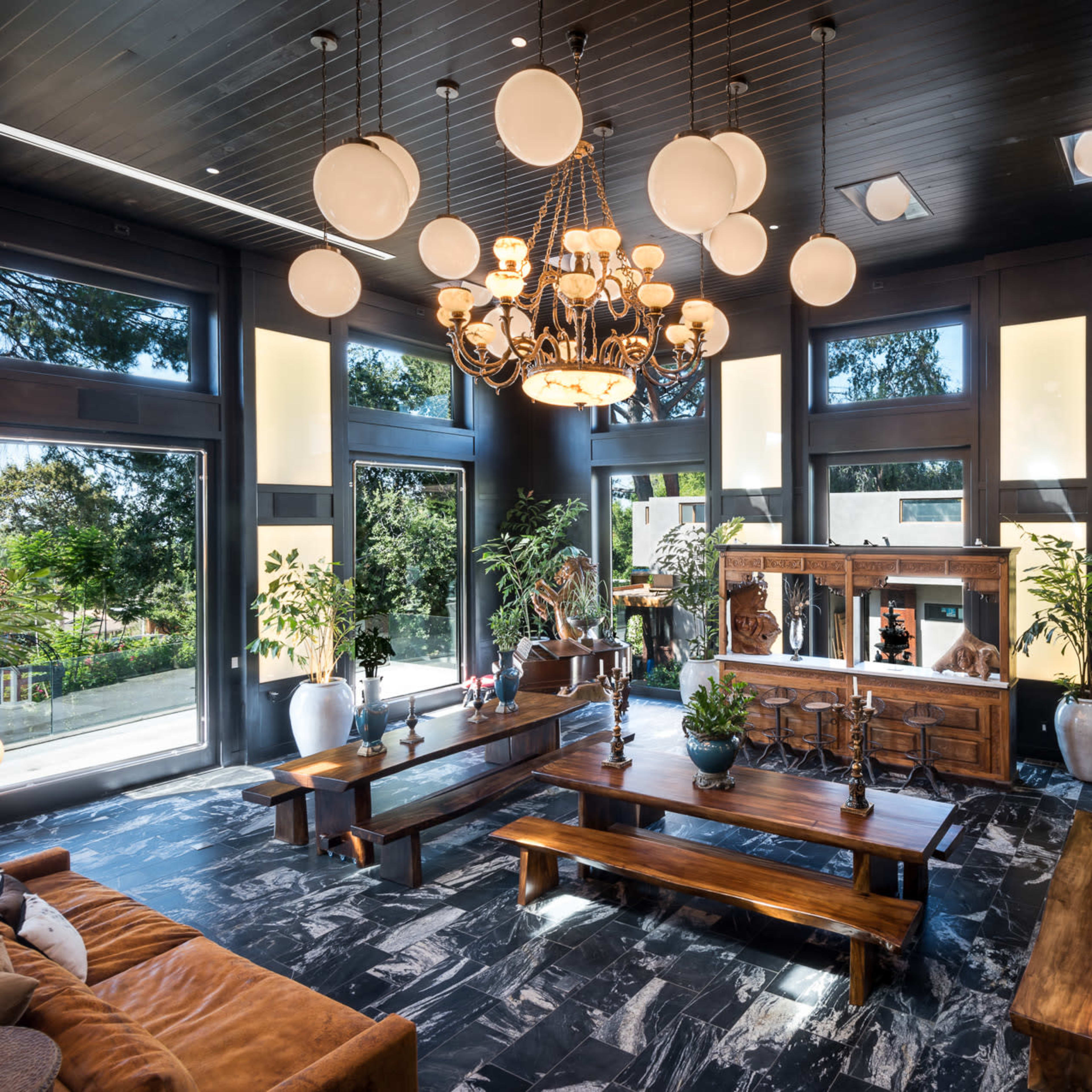 A spacious living room with large windows, a central chandelier, wooden tables, and decorative greenery.