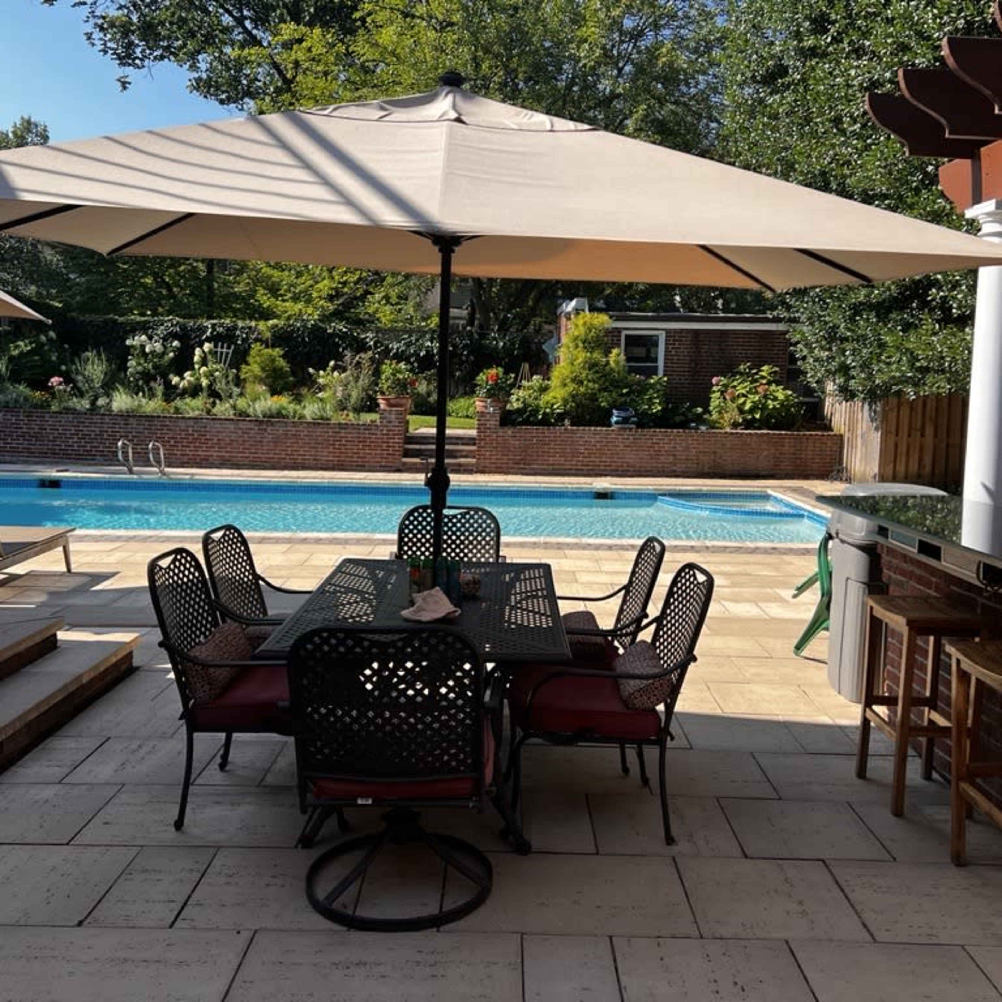 A patio area features a large dining table set with chairs, shaded by an umbrella, alongside a swimming pool surrounded by greenery.