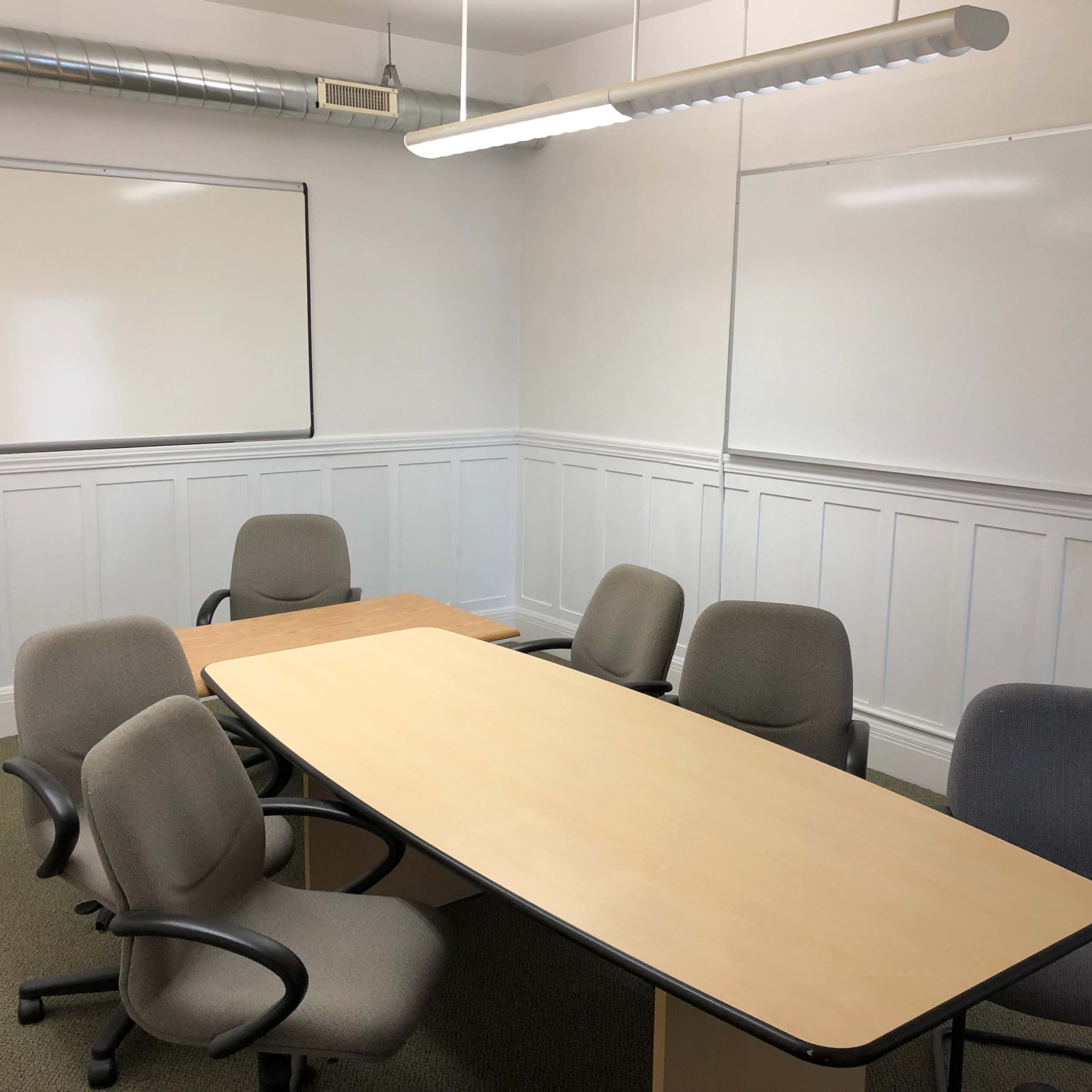 A small meeting room features an oval table surrounded by chairs, with two whiteboards on the walls.