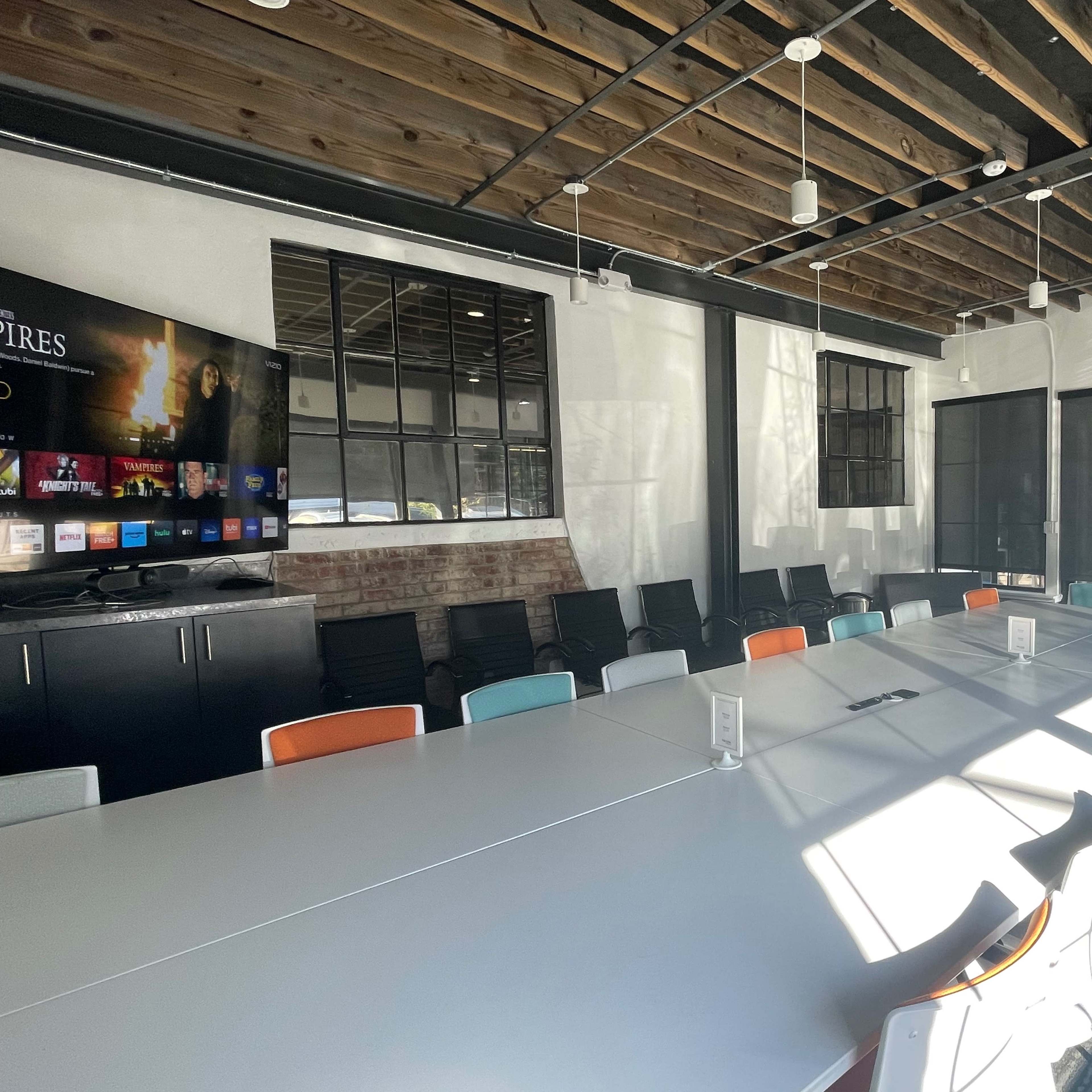 A modern conference room features a long rectangular table surrounded by chairs, with large windows and a wall-mounted screen displaying a menu.
