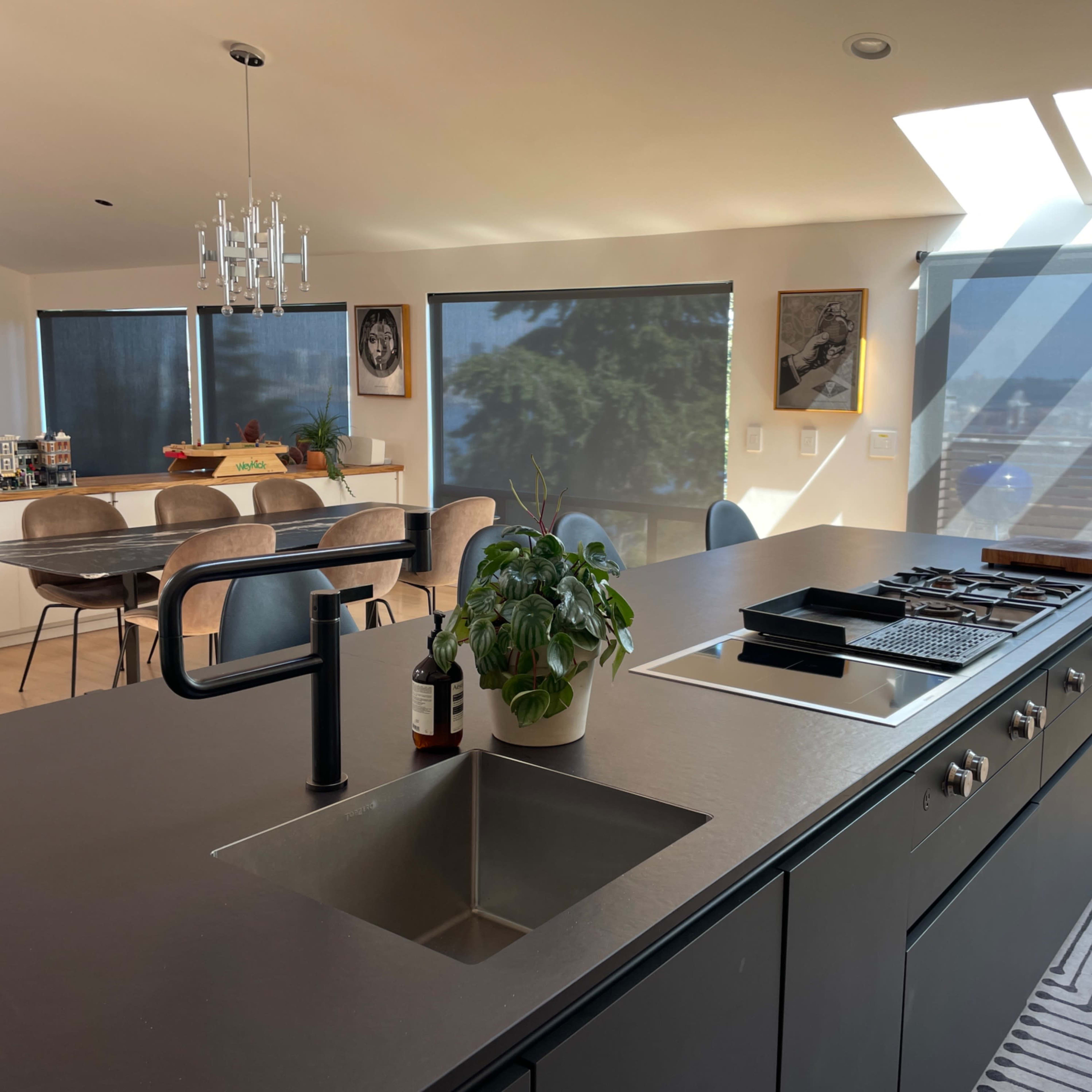 A modern kitchen with a central island featuring a sink and a cooktop, overlooking a bright dining area through large windows.