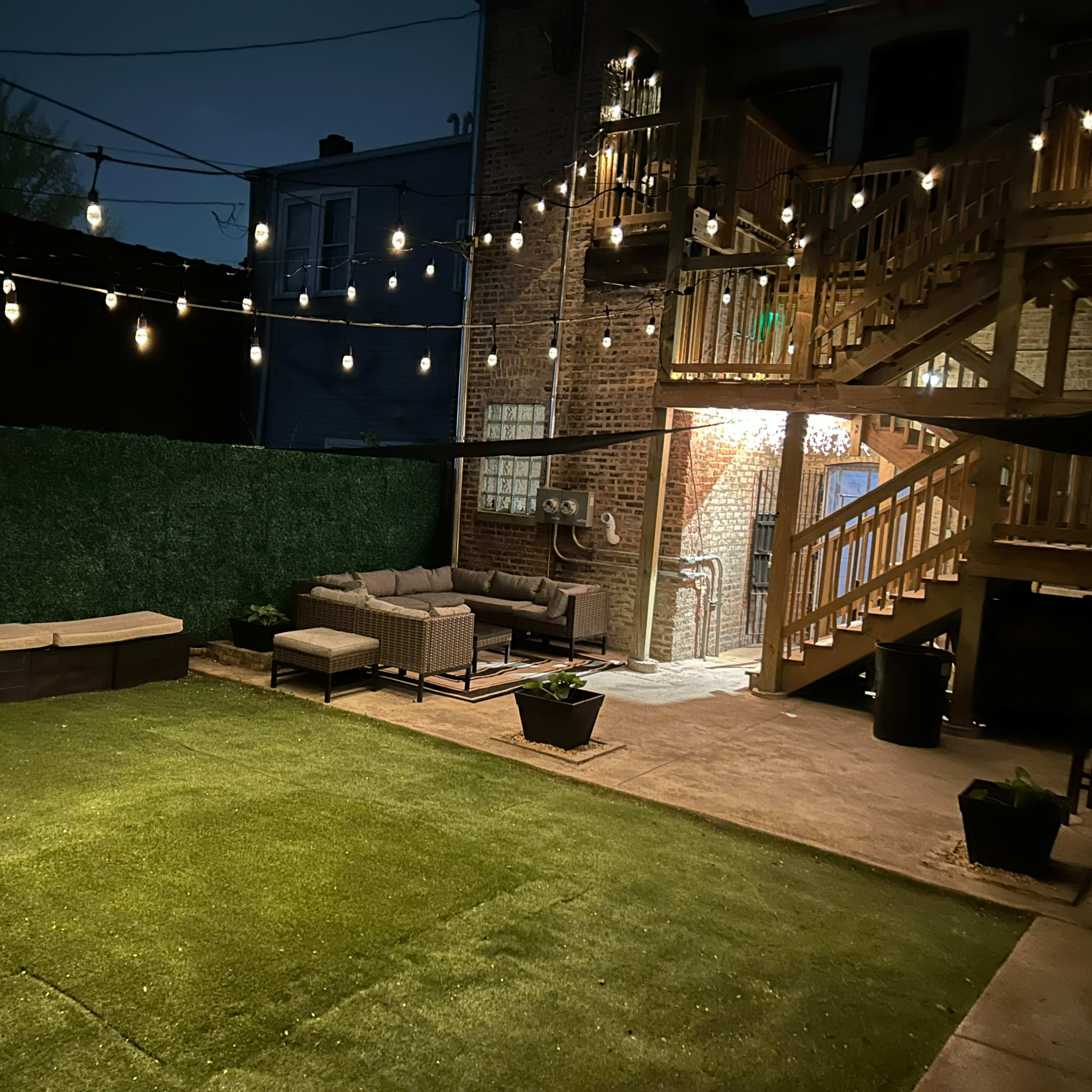 A dimly lit backyard with string lights, artificial grass, seating areas, and a staircase leading to an upper level.