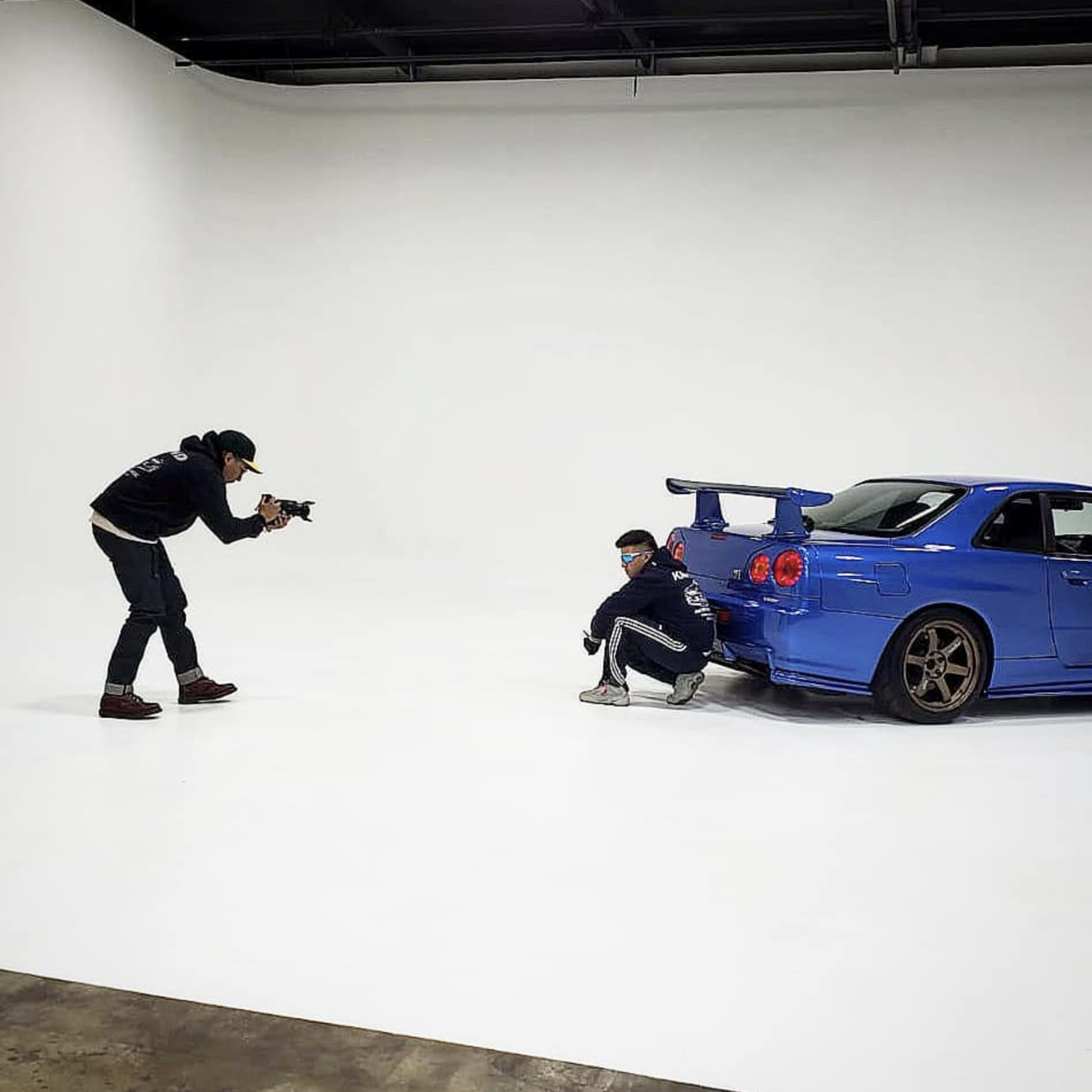 A photographer captures a posed shot of a person crouching next to a blue sports car in a studio setting with a white backdrop.