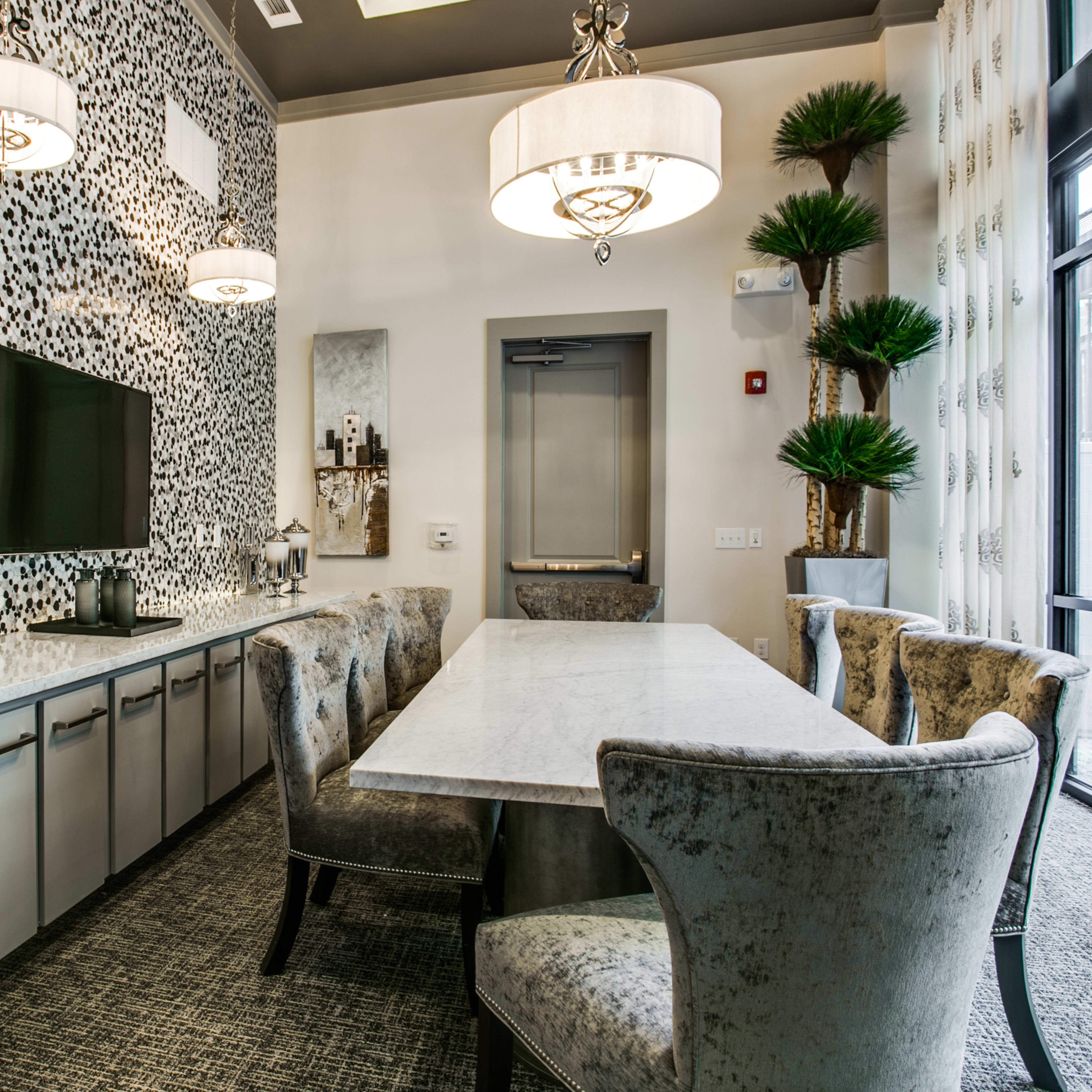 A modern dining area with a long marble table, plush chairs, a patterned accent wall, and a wall-mounted television.