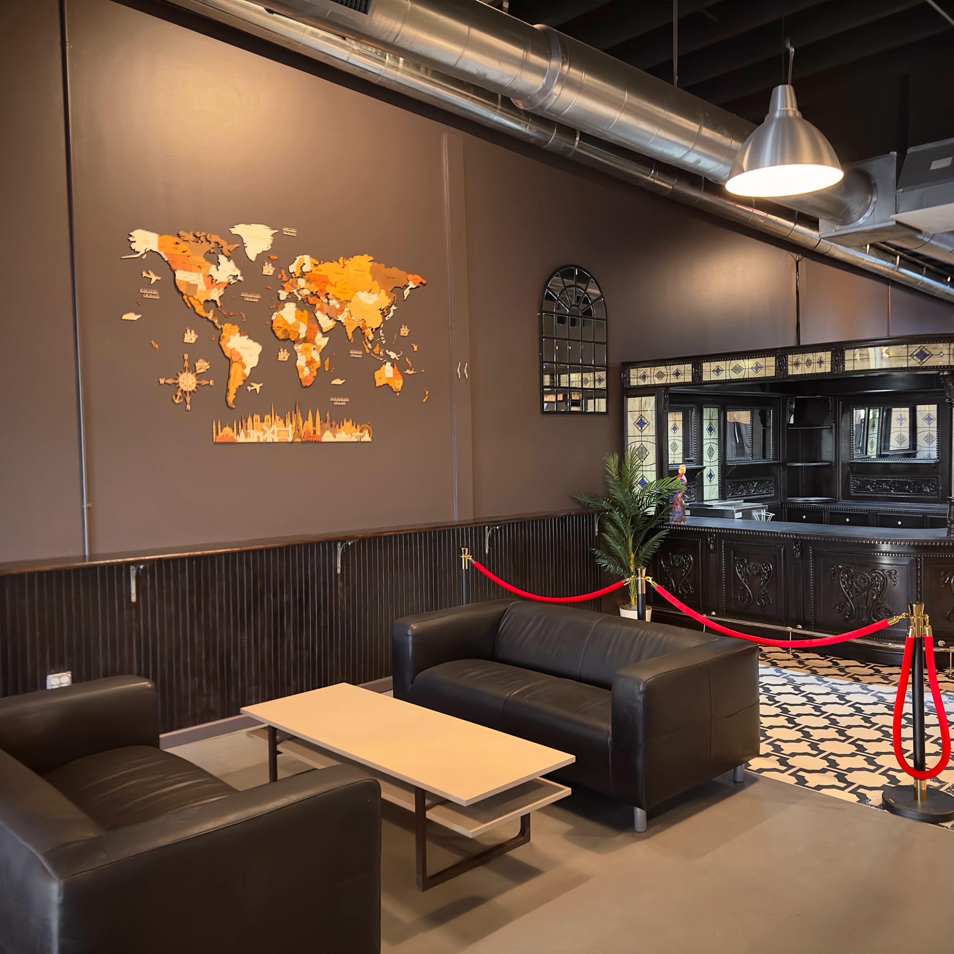 A modern interior space featuring two black sofas, a coffee table, a wall-mounted world map, and a bar area with a decorative backsplash, all set against dark walls and patterned flooring.