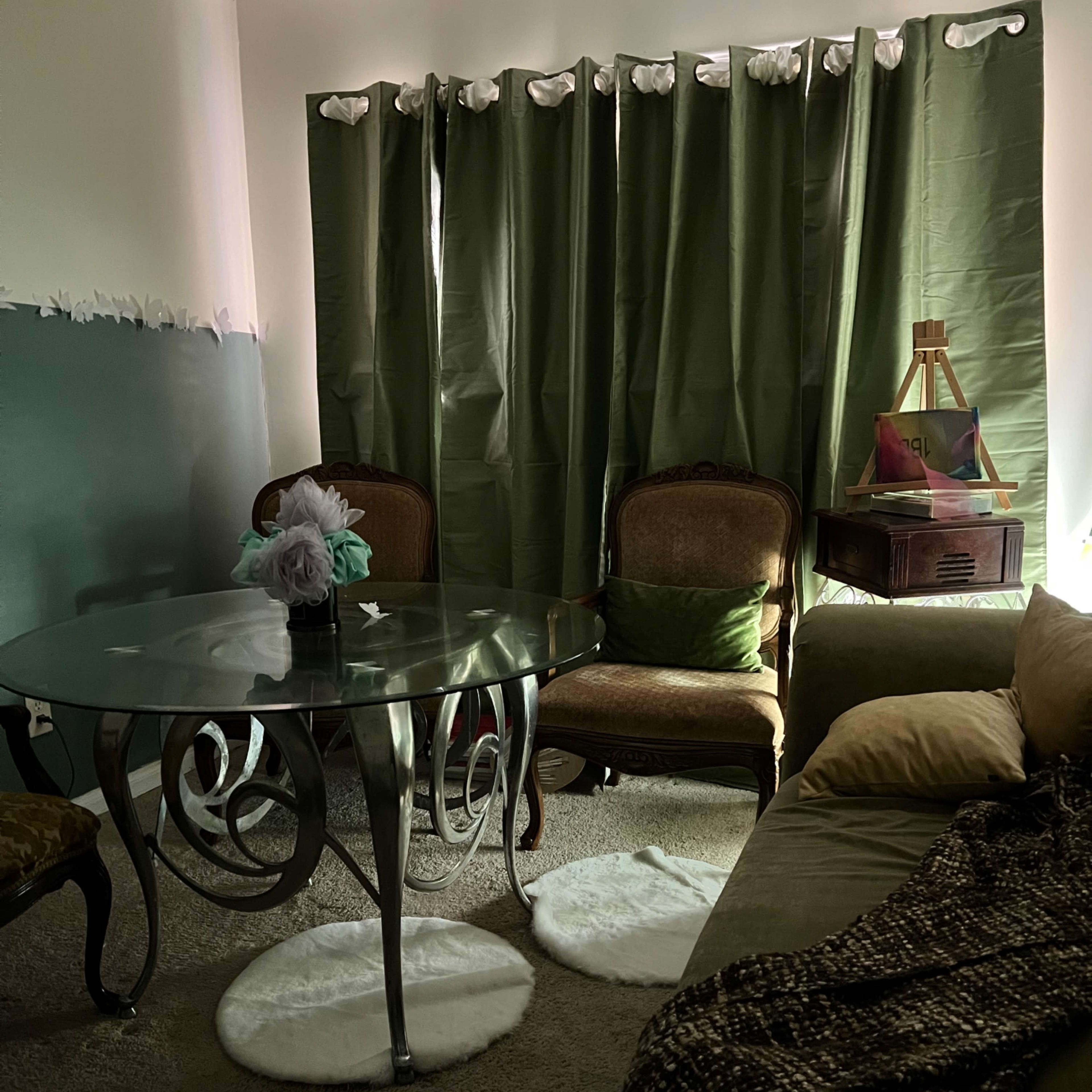 A living room features a glass dining table with curved legs, two upholstered chairs, a couch, and green curtains that block sunlight.