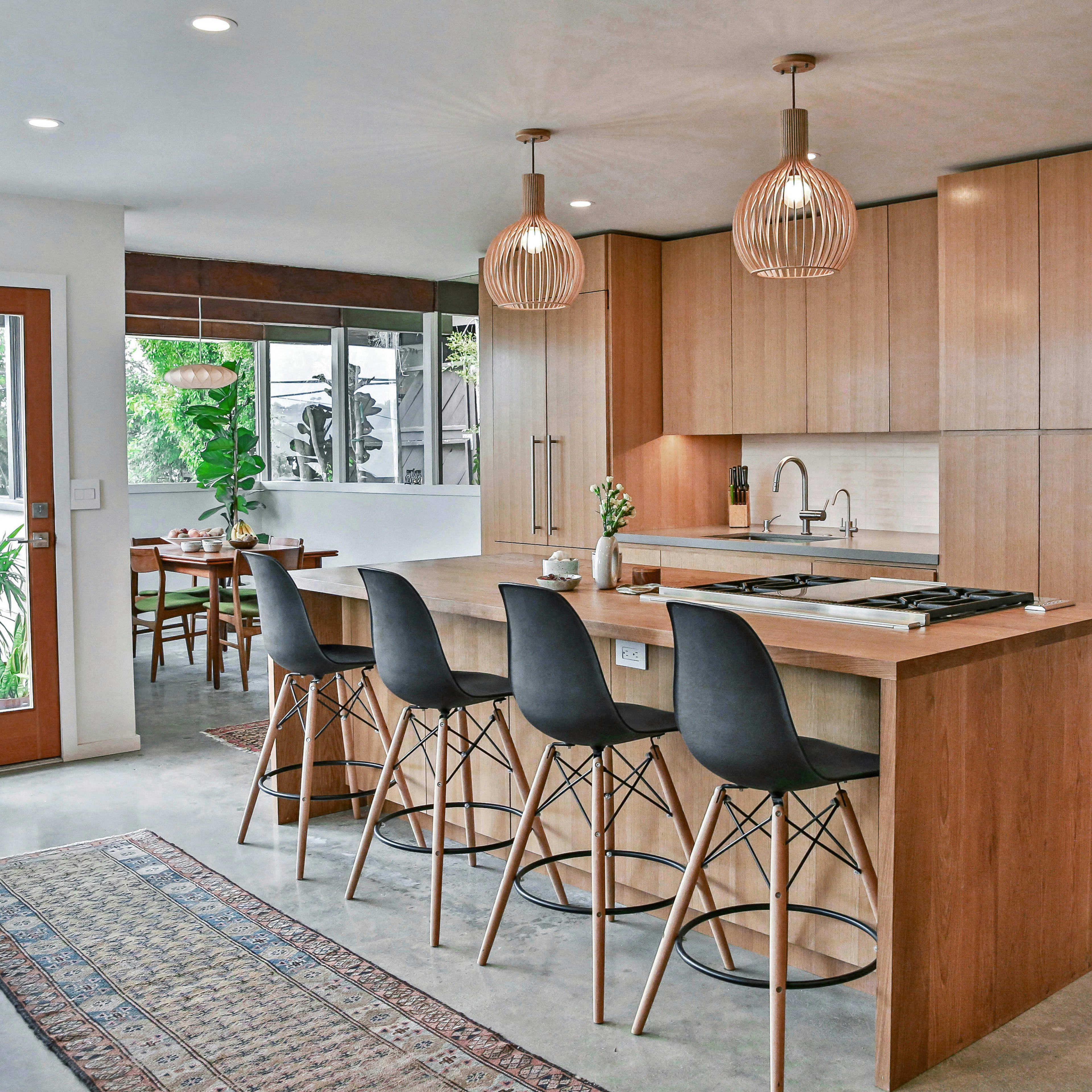 A modern kitchen features a wooden island with seating, pendant lights, and large windows overlooking a dining area.