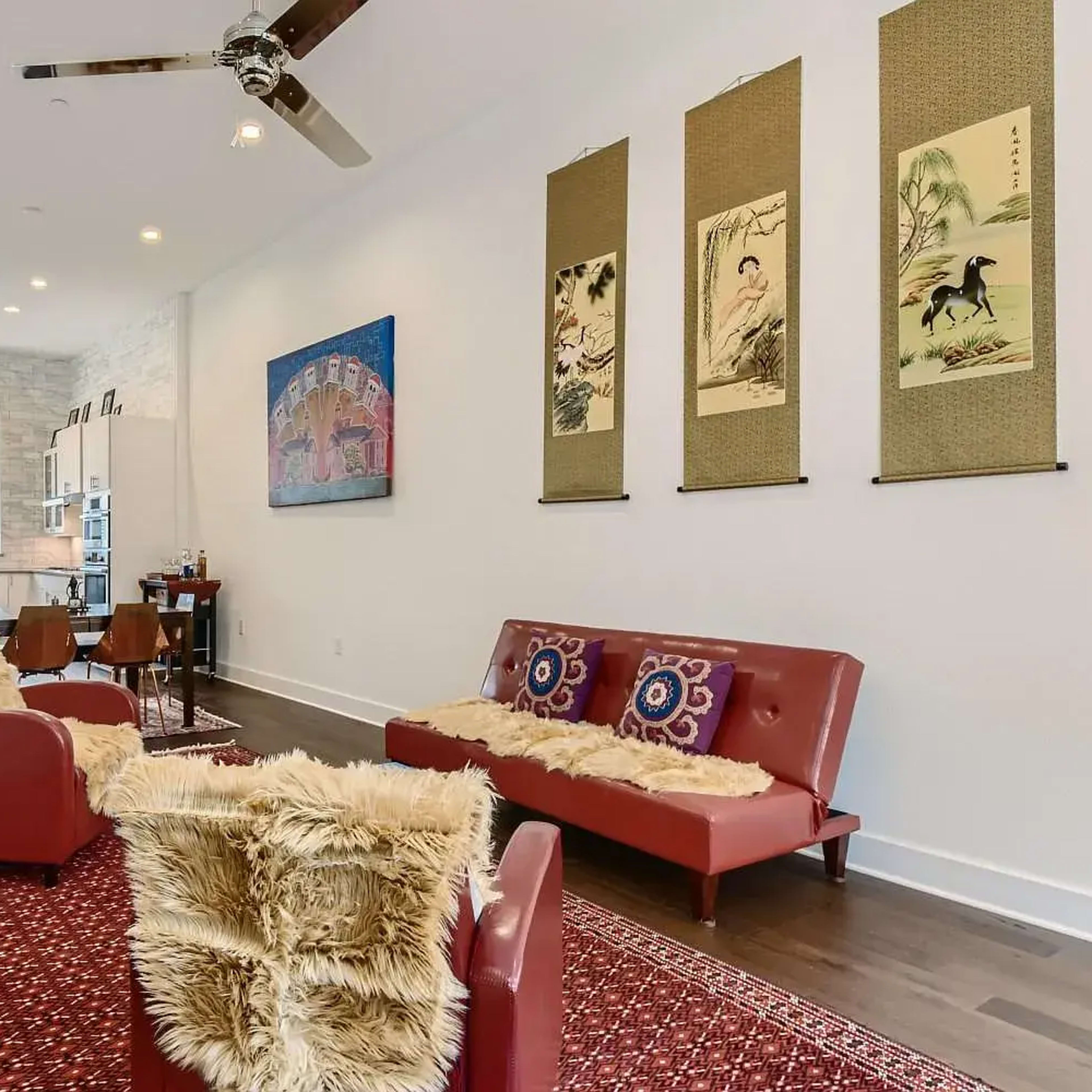A living room with red sofas, a patterned rug, and artwork on the walls, leading into a kitchen area visible in the background.