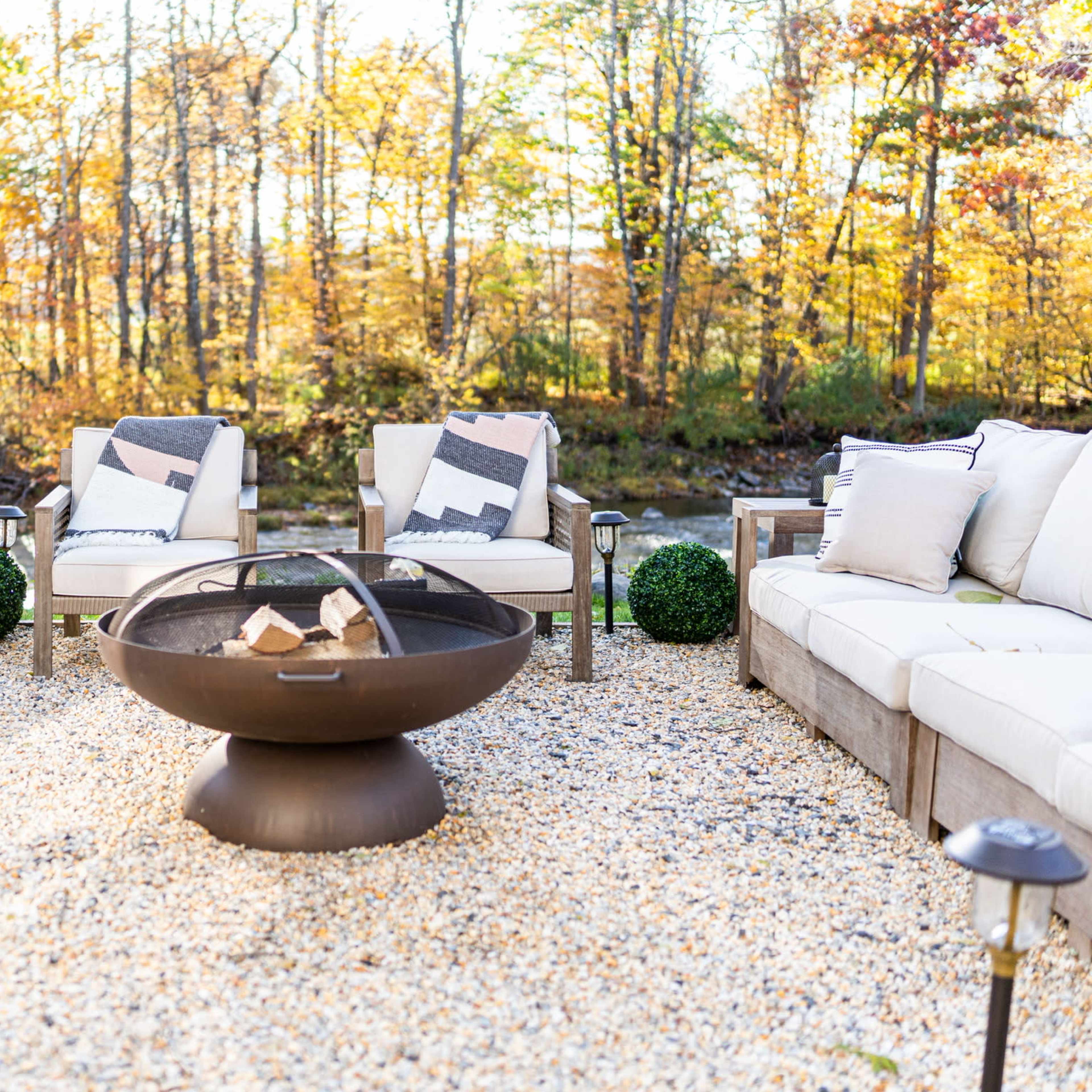A circular fire pit surrounded by an arrangement of outdoor seating and decorative plants in a gravel area, set against a backdrop of trees with autumn foliage.