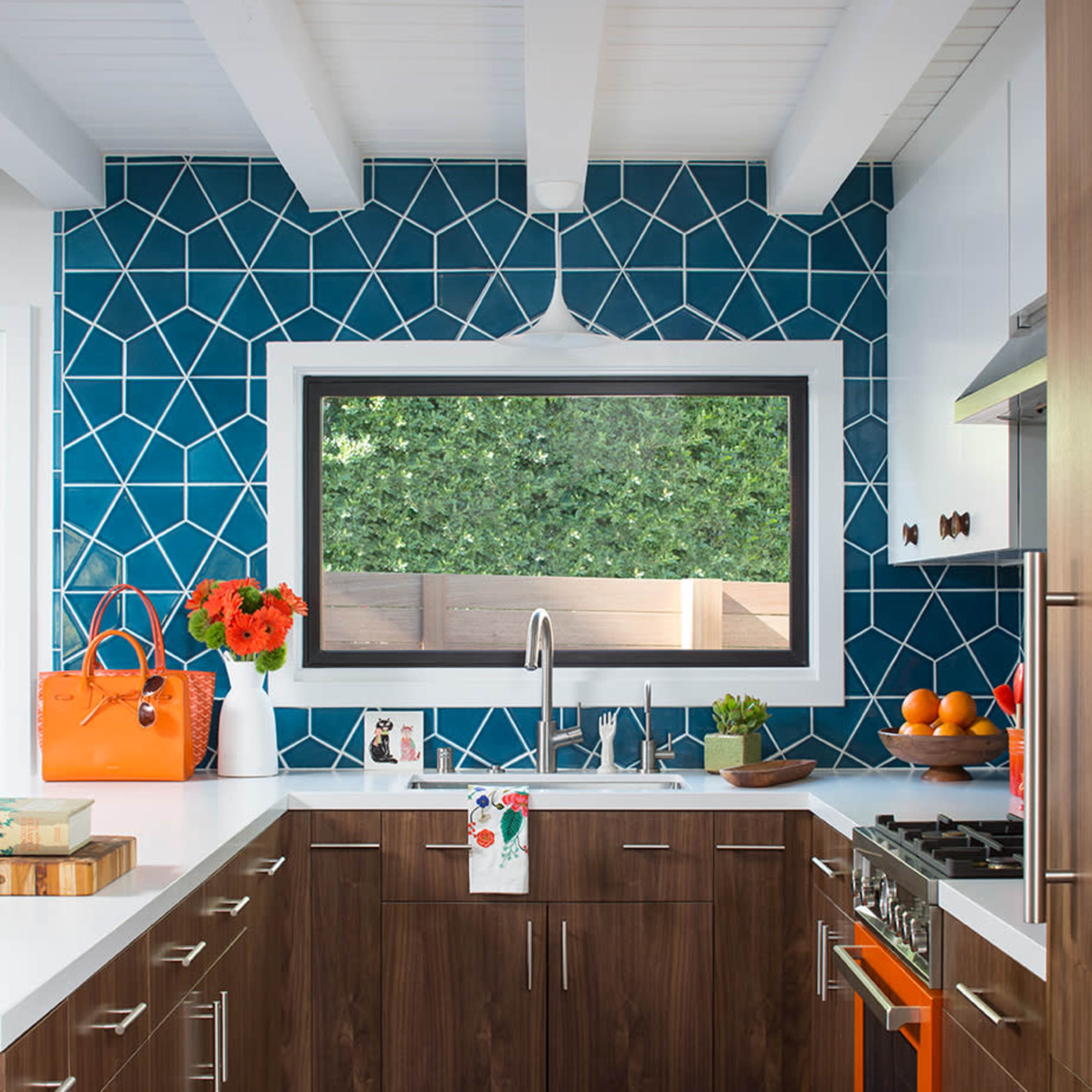 A modern kitchen features a bold geometric blue tile backsplash, wooden cabinetry, and orange accents, including an oven and a handbag.