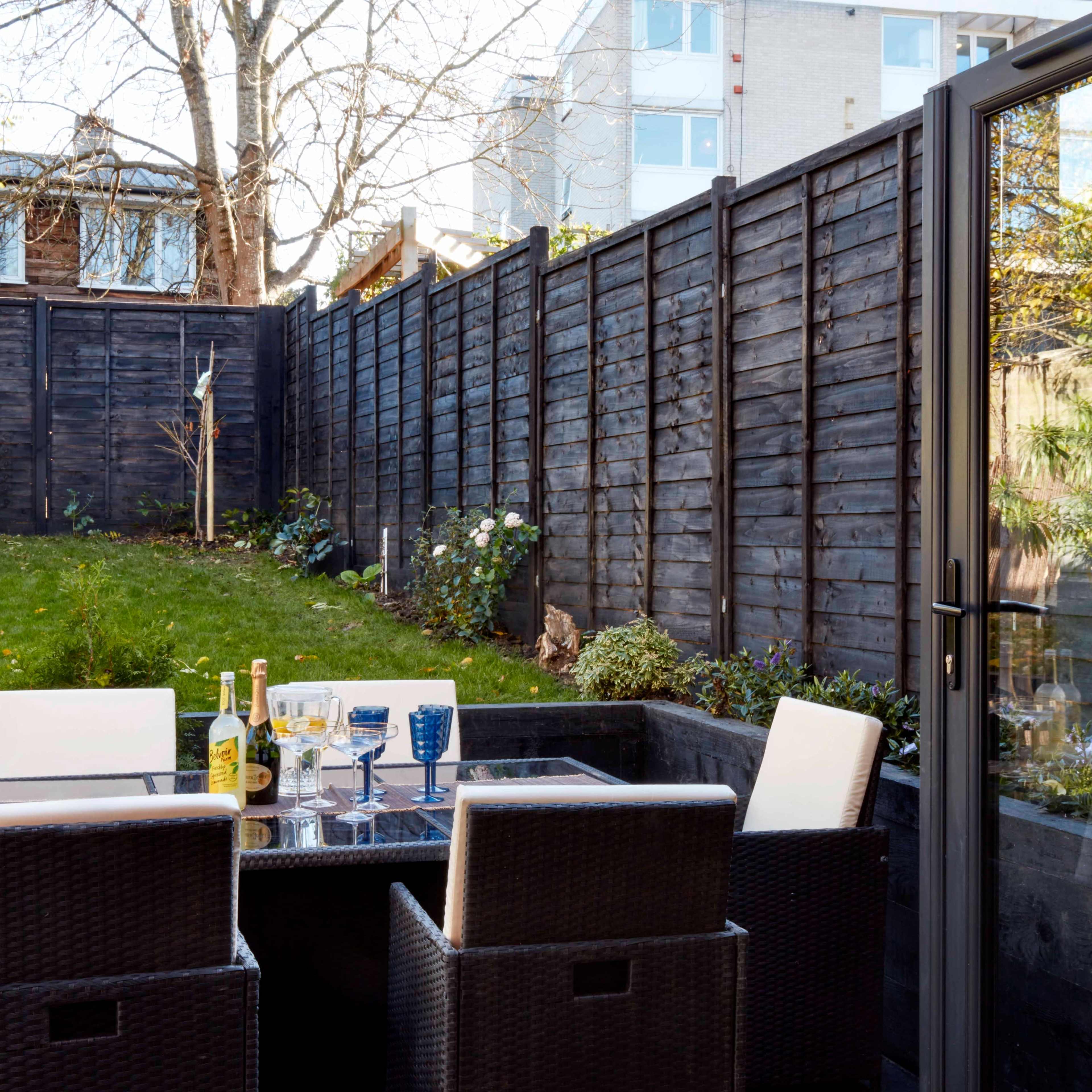 A patio area features a glass-top table surrounded by wicker chairs, with a view of a well-maintained garden enclosed by wooden fencing.