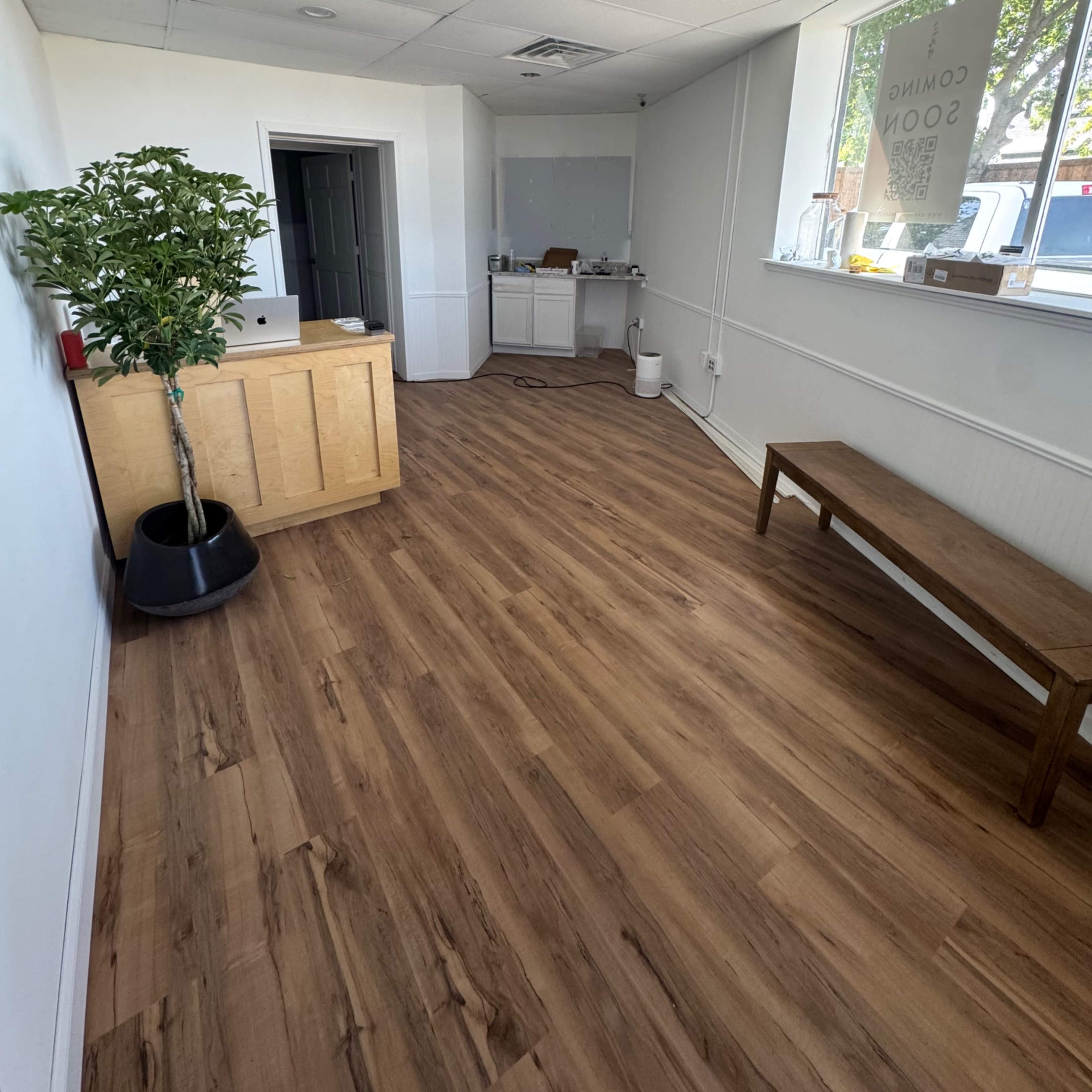 An empty office space with wooden flooring, a reception desk, a bench, and a large window.