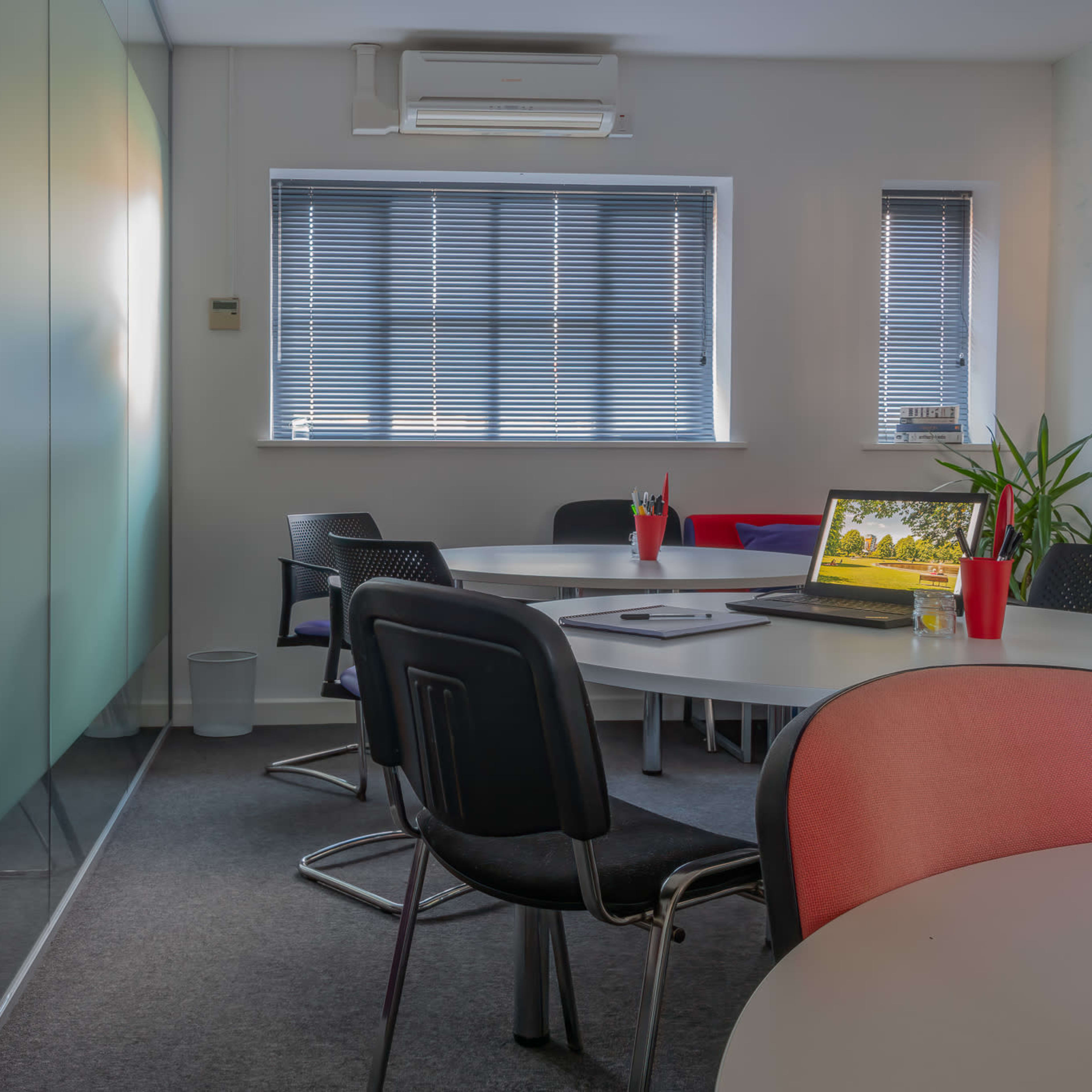 A modern conference room features a round table surrounded by black and red chairs, with a laptop open on the table and large windows partially covered by blinds.
