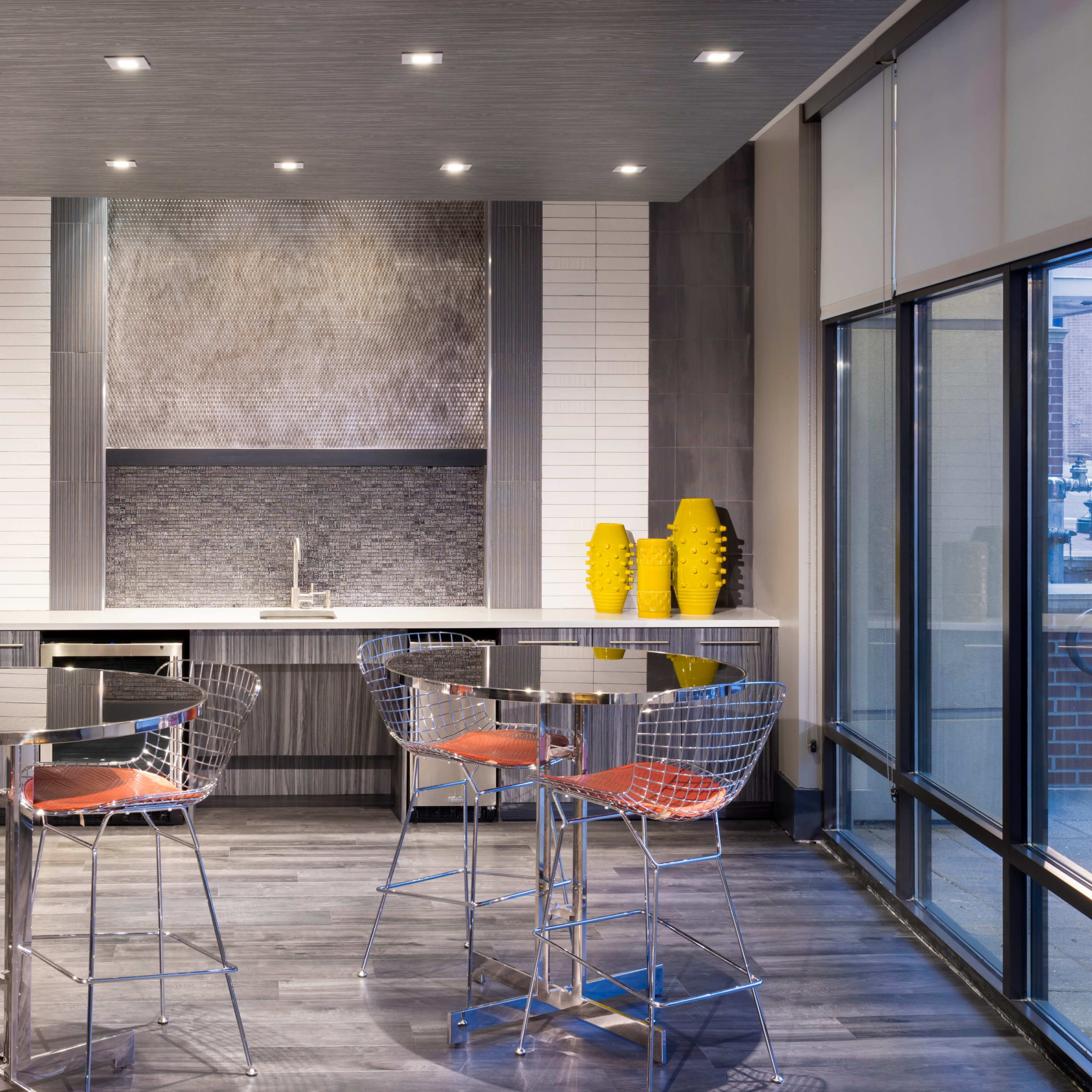 A modern kitchen area with a bar-style dining set, featuring chrome stools and a sleek countertop, complemented by decorative yellow vases and wall art.