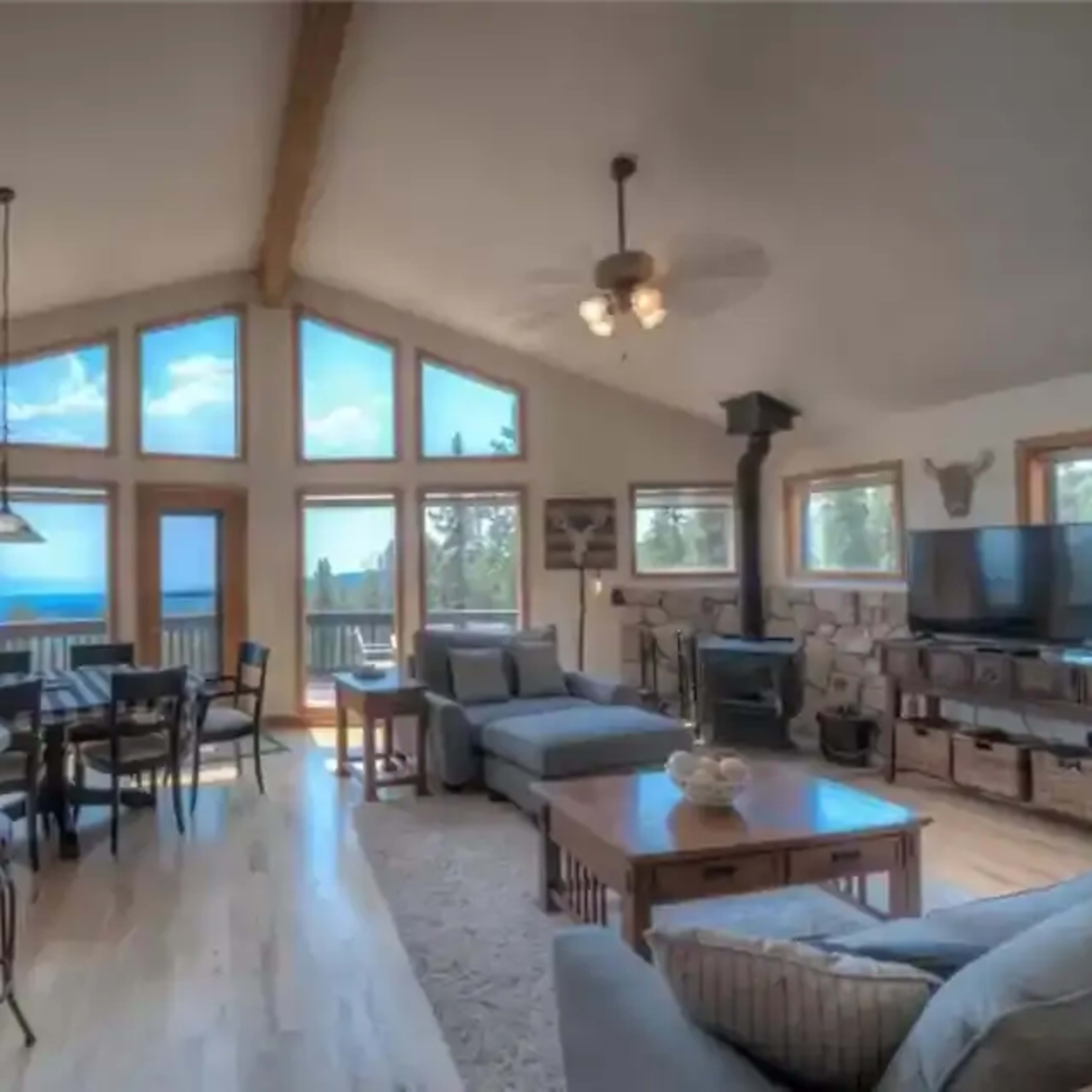 A spacious living area with large windows offering a view of the outdoors, featuring a dining table, kitchen, and comfortable seating.